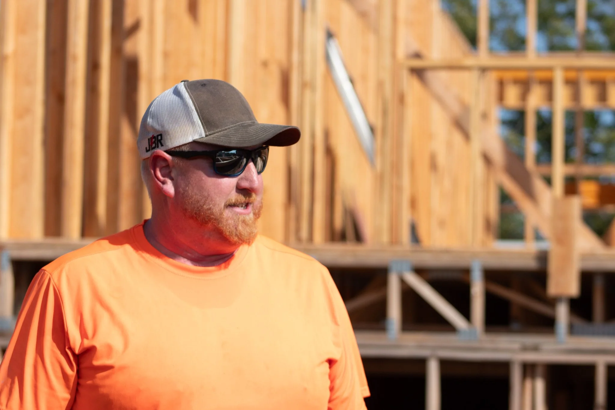 A man in sunglasses, a baseball cap, and an orange shirt standing in front of a wooden construction site.