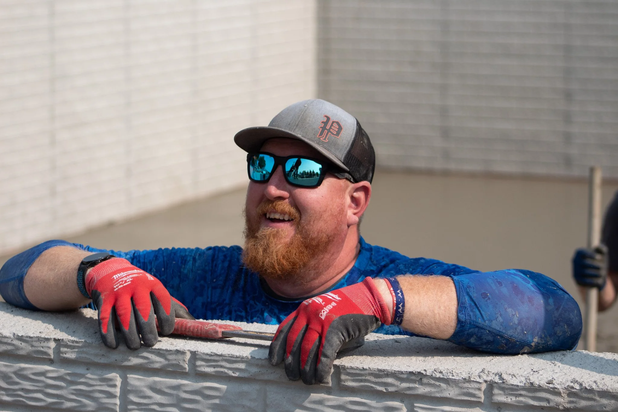 Man with a beard wearing sunglasses, a gray cap, red gloves, and a blue shirt, stretching his arms over a white concrete wall.