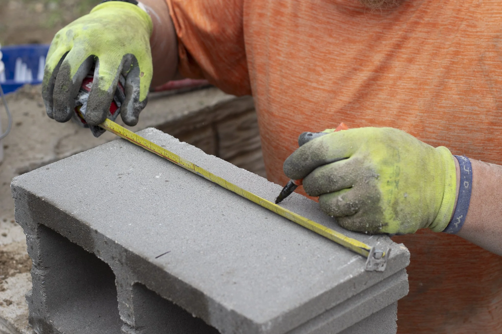 A person wearing yellow work gloves and an orange shirt is using a measuring tape and marker to mark a concrete block on a construction site. The scene shows a close-up of their hands and the gray concrete block, with a blurred background.