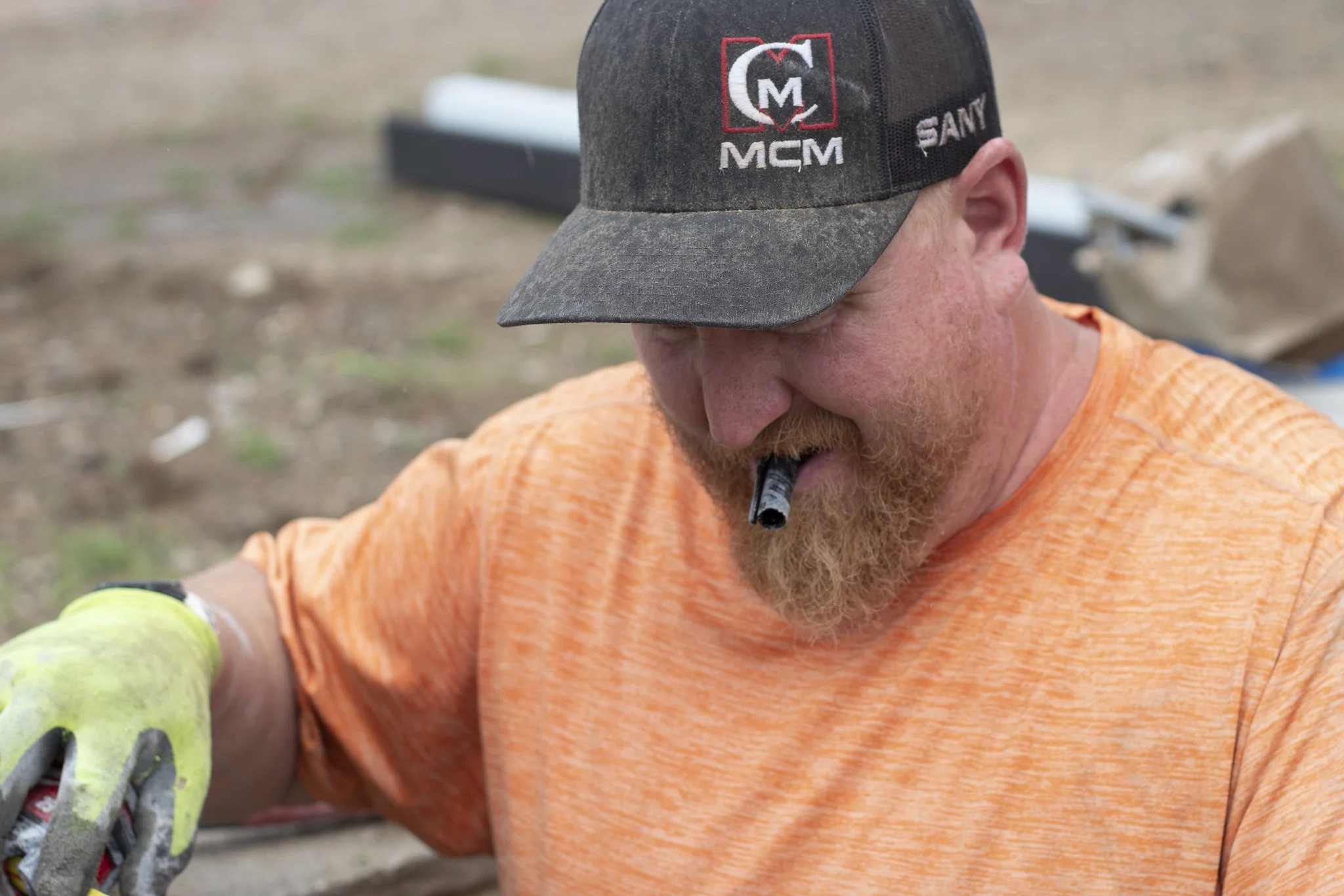 A man with a red beard wearing an orange shirt, a gray cap with the logo 'MCM' and the word 'SAM', and a neon yellow glove, working outdoors on a construction or landscaping project.