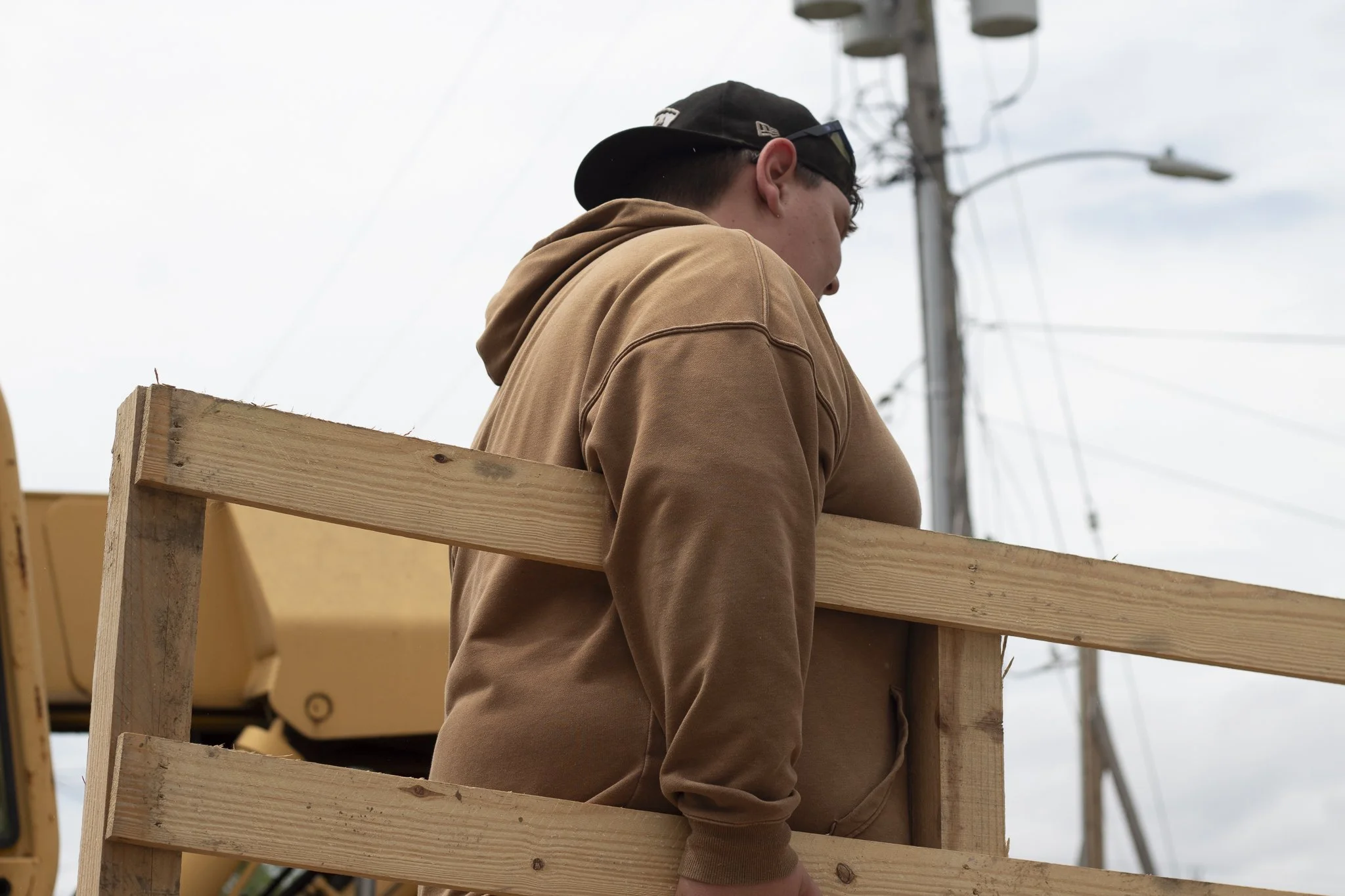 A person wearing a brown hoodie and black cap standing on a wooden platform outdoors with power lines in the background.