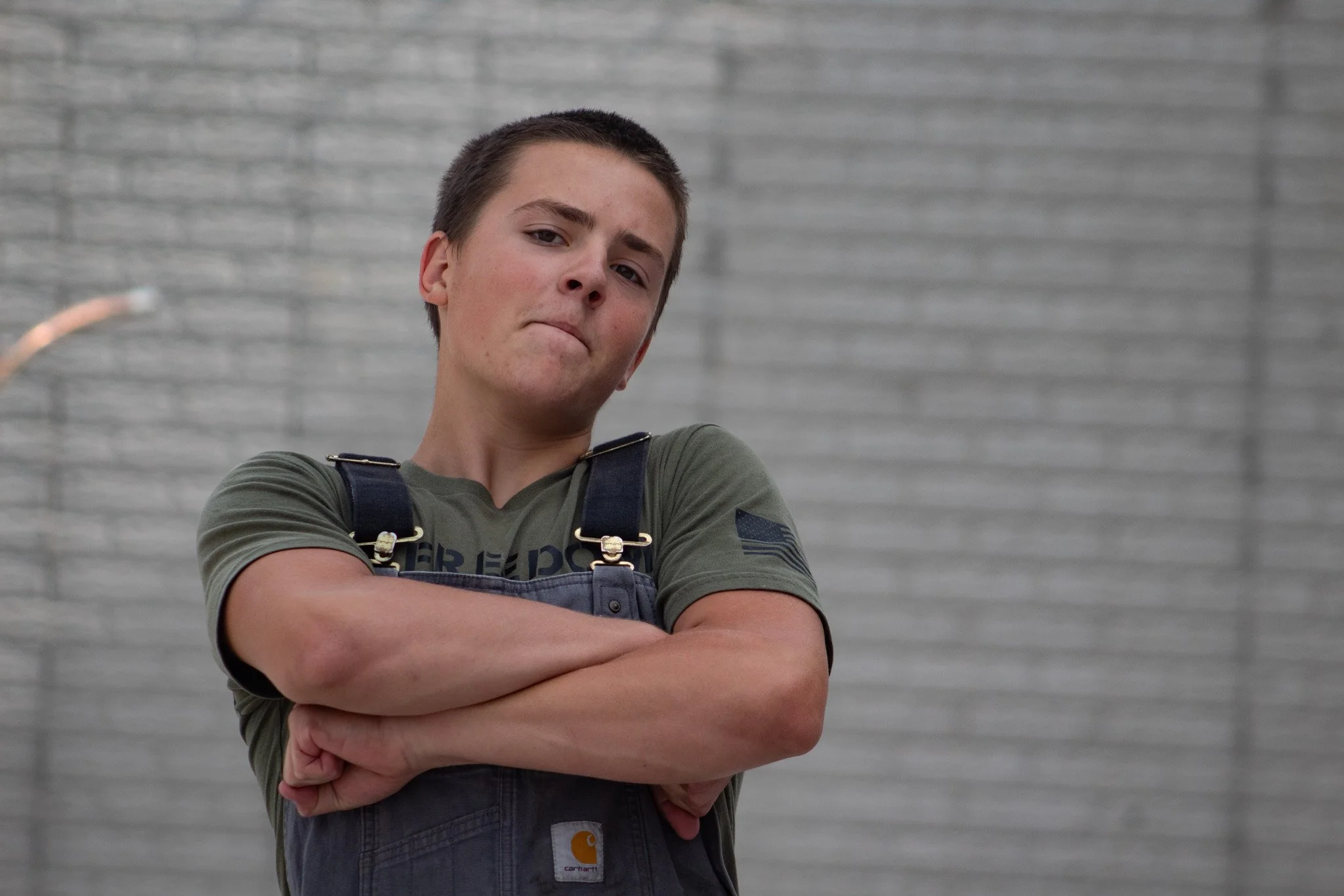 A young man with short brown hair, crossed arms, and confident expression standing against a gray brick wall. He wears a green T-shirt and overalls.