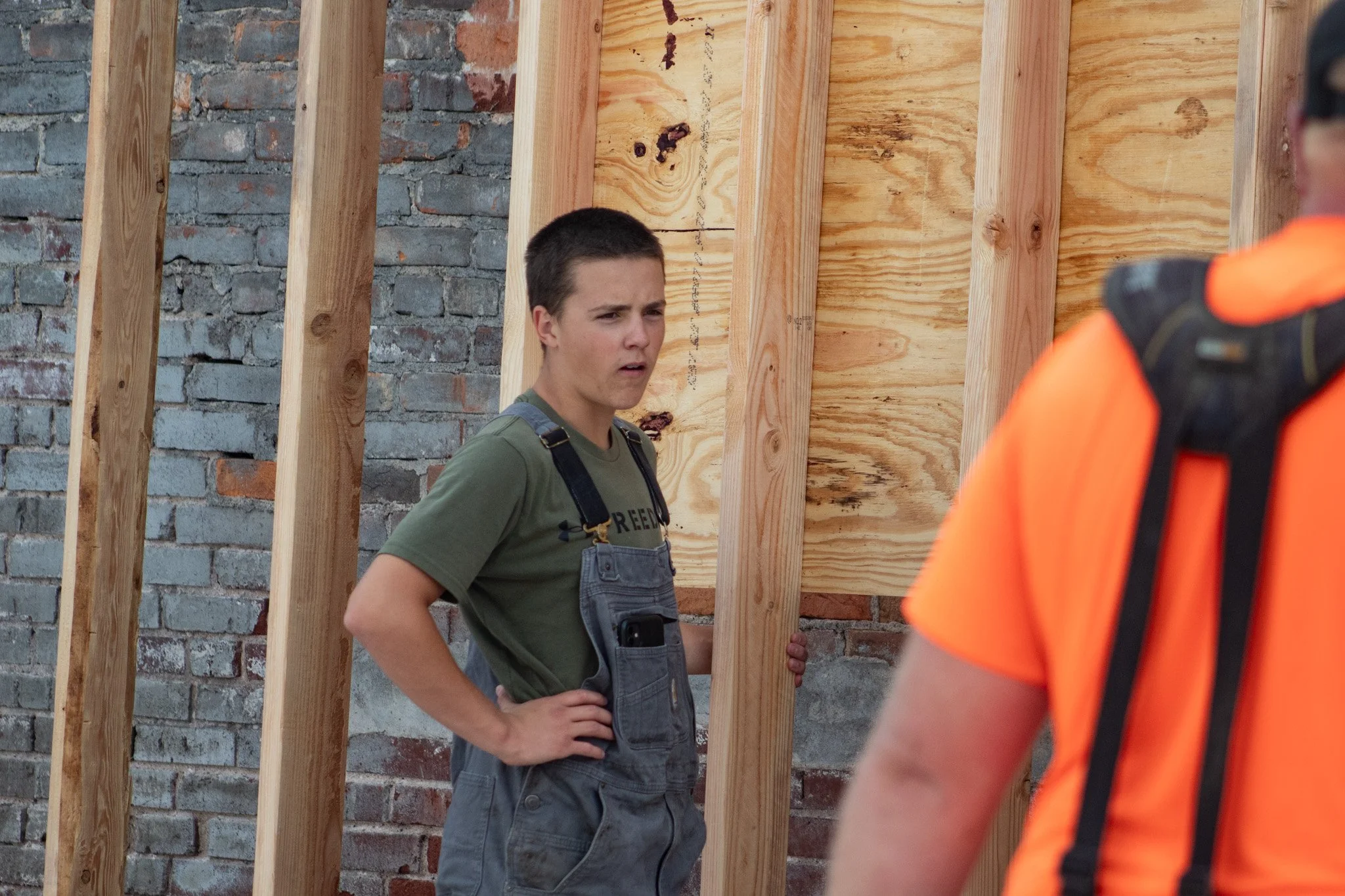 A young man wearing work overalls and a green T-shirt standing on a construction site with wooden framing and an unfinished brick wall behind him, looking at a person in an orange safety vest.