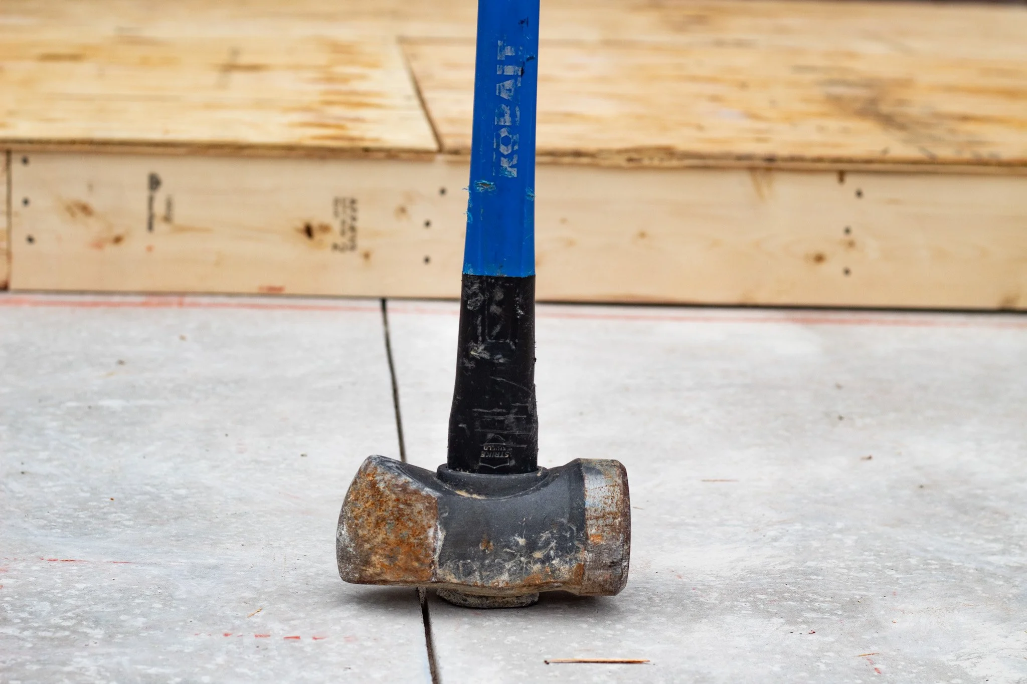 A close-up of a small hammer with a rusted head, a blue handle, and a black grip, standing upright on a concrete surface with a wooden wall in the background.