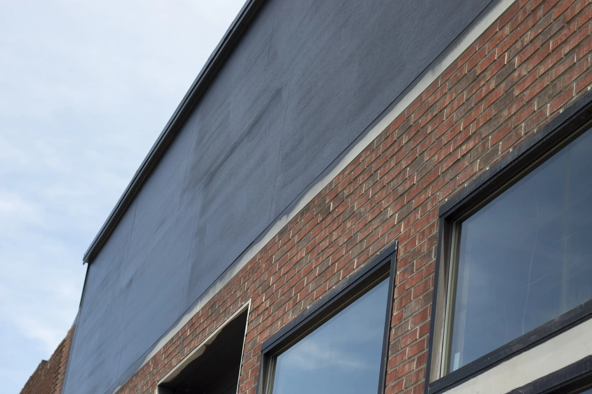 Close-up view of a modern building with a corner showing red brick wall and large windows, part of a gray building exterior, under a blue sky.