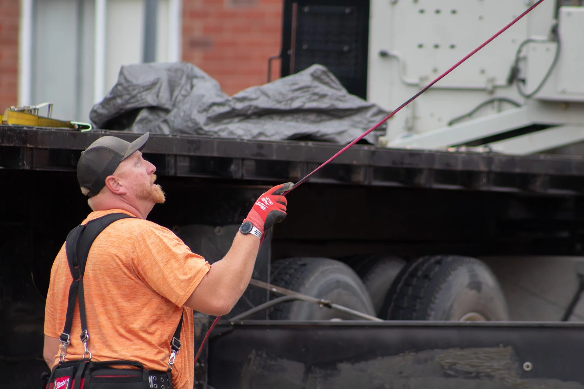 A man with a beard and cap, wearing an orange shirt and red gloves, is working with tools near a large truck. He appears to be engaged in some maintenance or repair work.