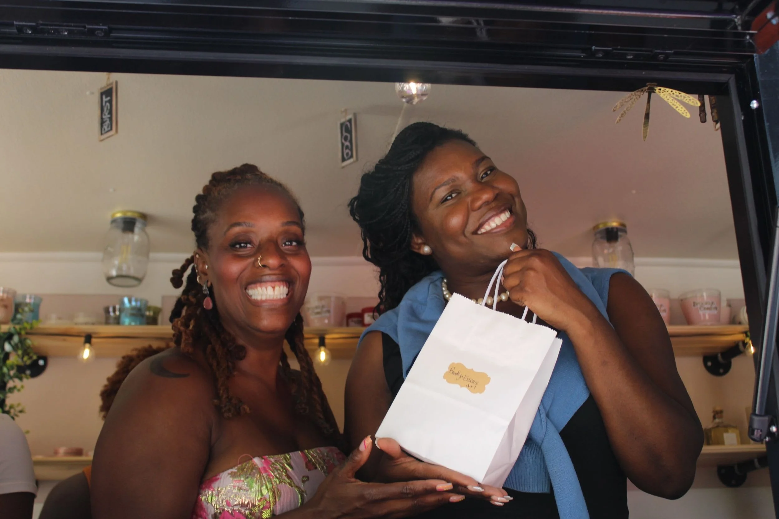 Two smiling women standing in a shop, one holding a gift bag, with shelves and decorations in the background.