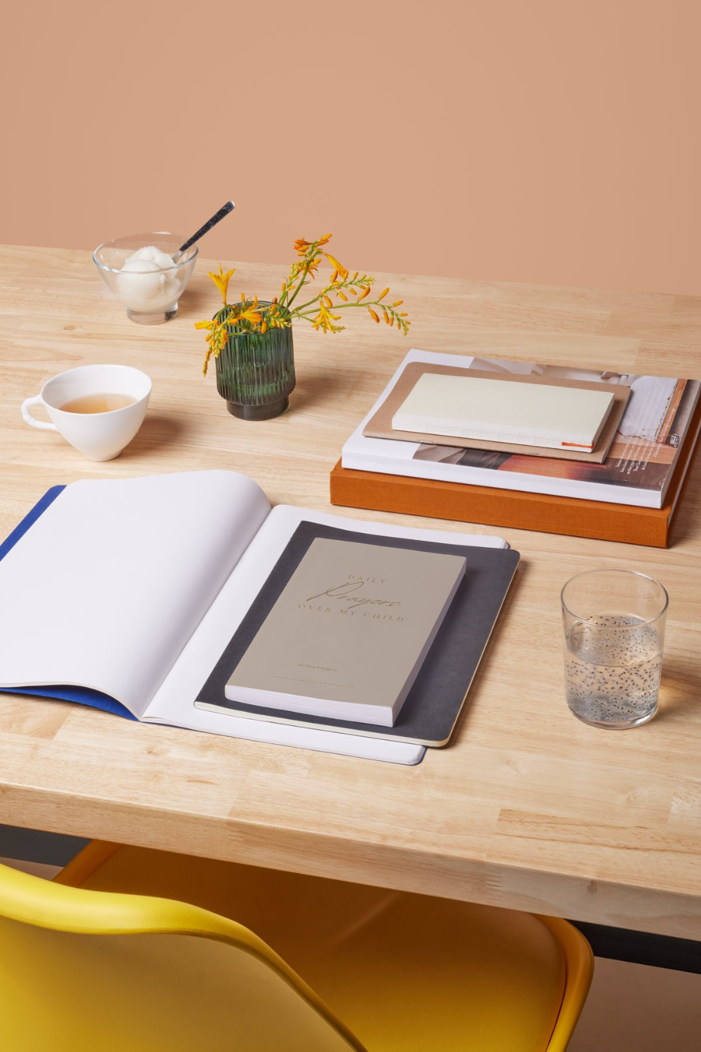 A wooden table with a yellow chair, an open notebook, a closed book, a small potted plant with yellow flowers, a cup of tea, a glass of water, a bowl of ice cream with a spoon, and a stack of magazines.