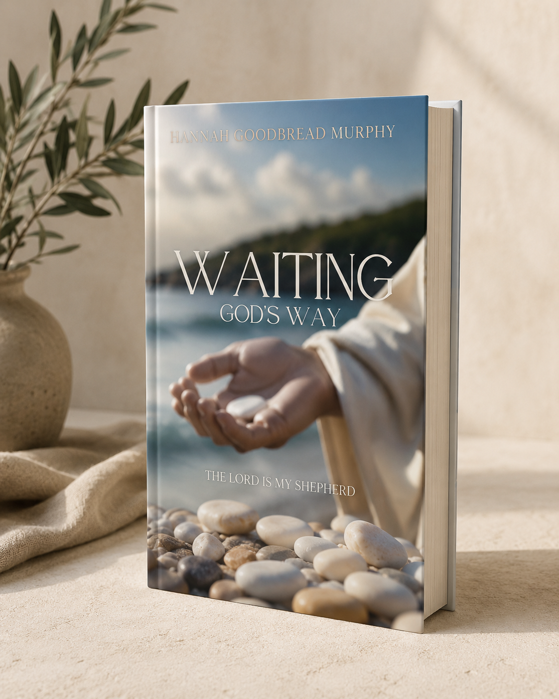 A book titled "Waiting God's Way" by Hannah Goodbread Murphy, featuring a photo of a hand holding a small stone, with a beach and ocean in the background, and a Bible verse 'The Lord is my shepherd.'