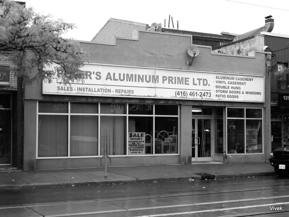 A storefront with a sign for R's Aluminum Prime Ltd., advertising sales, installation, and repairs of aluminum products like casements, vinyl casement, double hung, storm doors and windows, and patio doors. The window displays a sale sign offering 50% off, with the store's phone number.