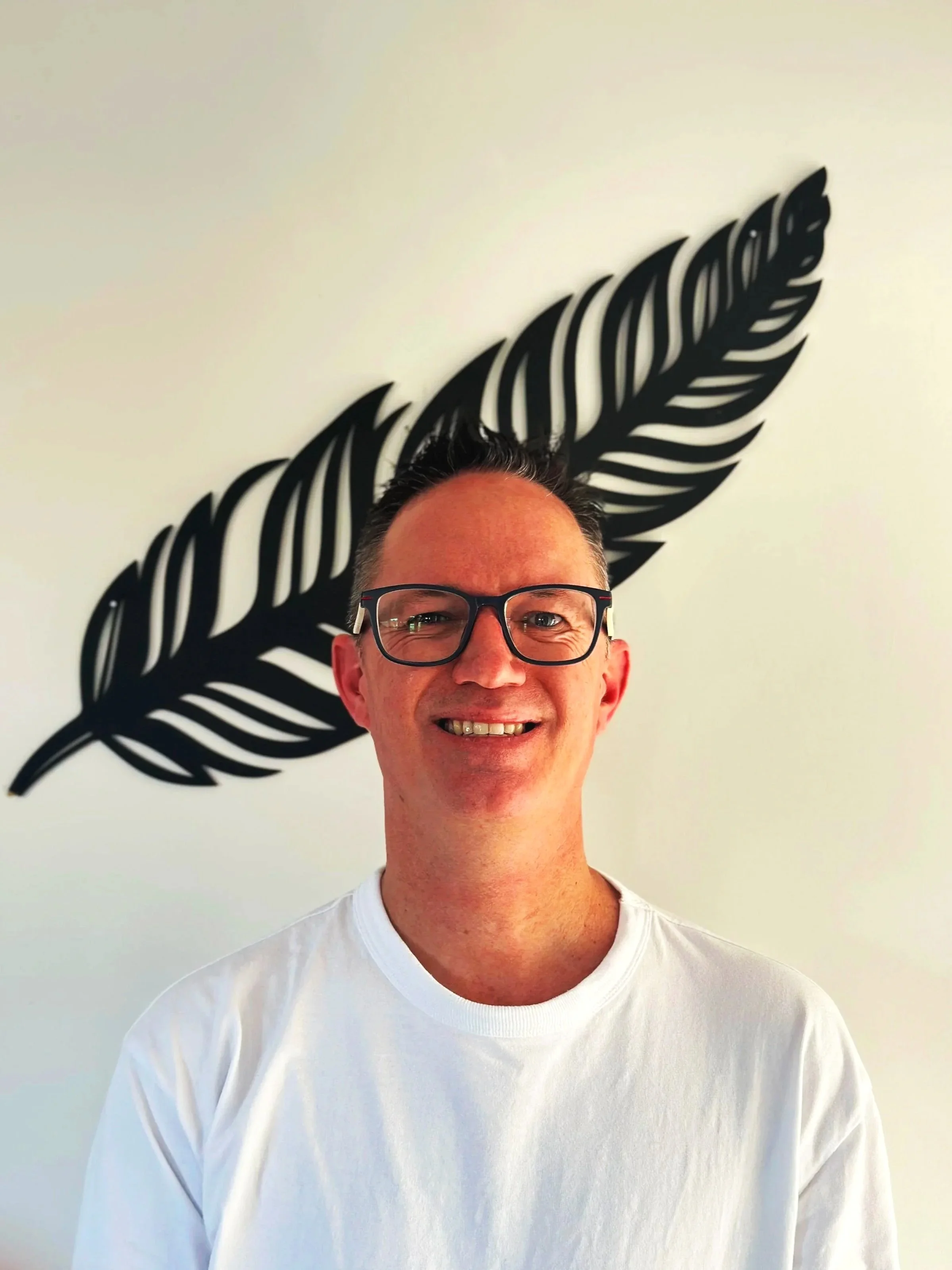 Smiling man with glasses in front of a white wall with a black decorative leaf design.