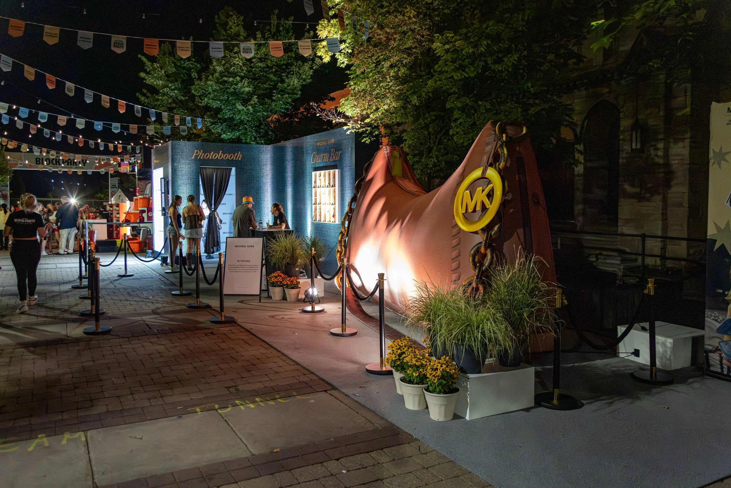 Night scene of an outdoor event with people gathered near a blue photo booth labeled "Photobooth" and a charm bar, illuminated by lights. There are decorative banners hanging above and large digital screens visible. A giant pink shoe with a yellow Mi
