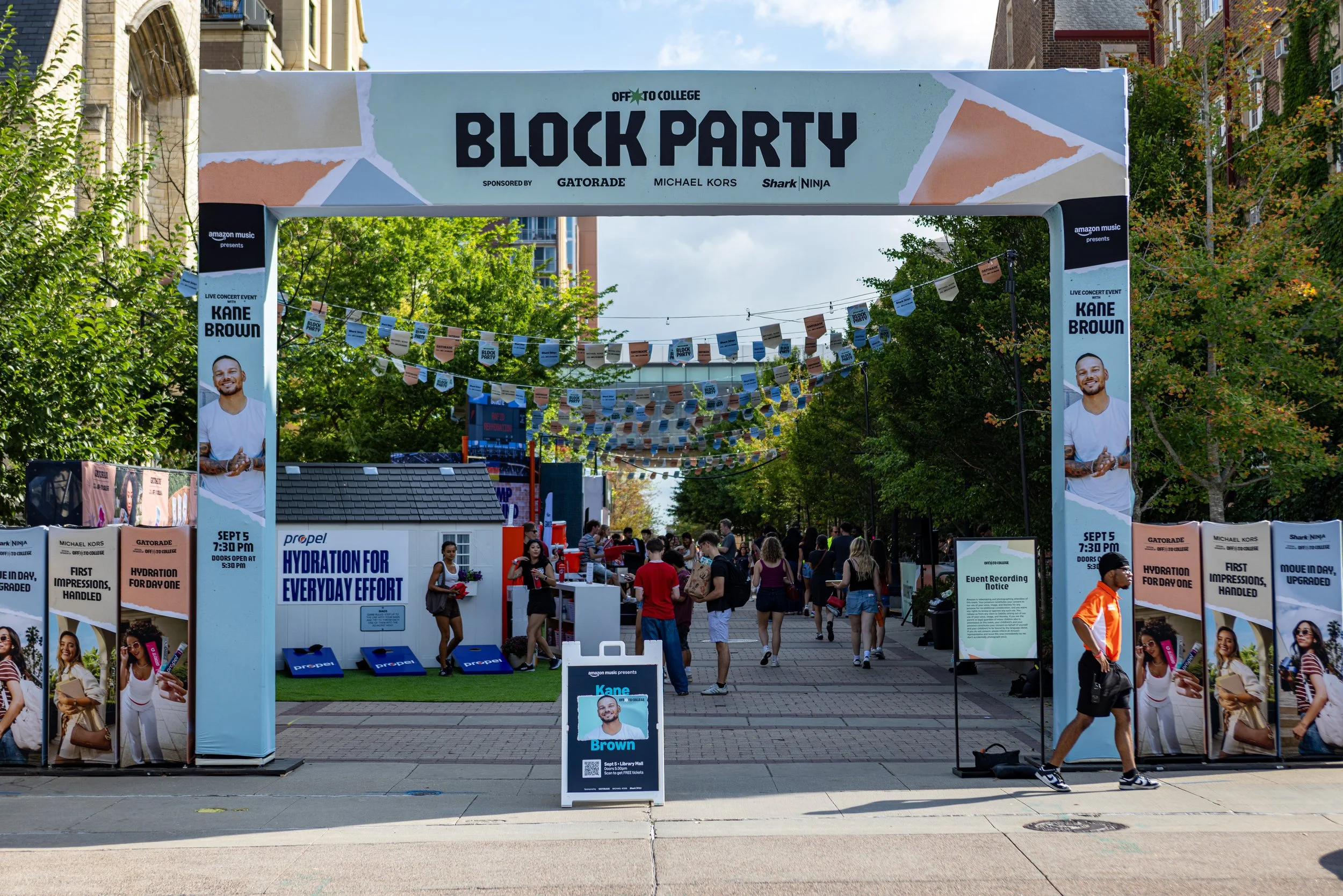 A street festival with a large archway entrance that reads 'Block Party' and features advertisements for an event with Kanye Brown scheduled for September 5th at 7:30 PM. The street is decorated with banners and string lights, with many people walkin