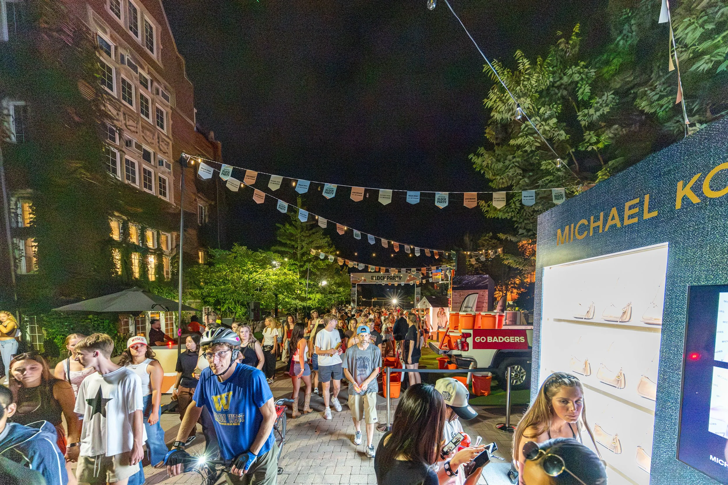 Crowd of young people at an outdoor night event with flags and banners overhead, some individuals looking at their phones, as a food or vendor stand labeled 'GO BADGERS' is visible in the foreground, and a large illuminated sign with portions of the 