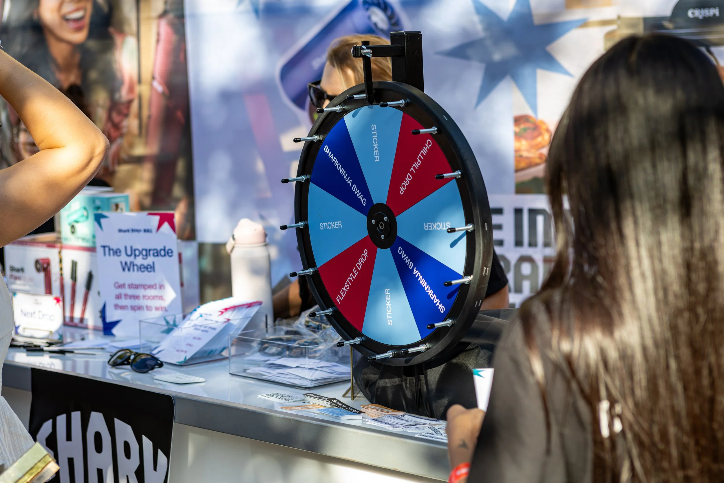 A spinning wheel with various prize options at a booth, including stickers, and a sign that reads "The Upgrade Wheel." People are gathered around, participating in the activity.