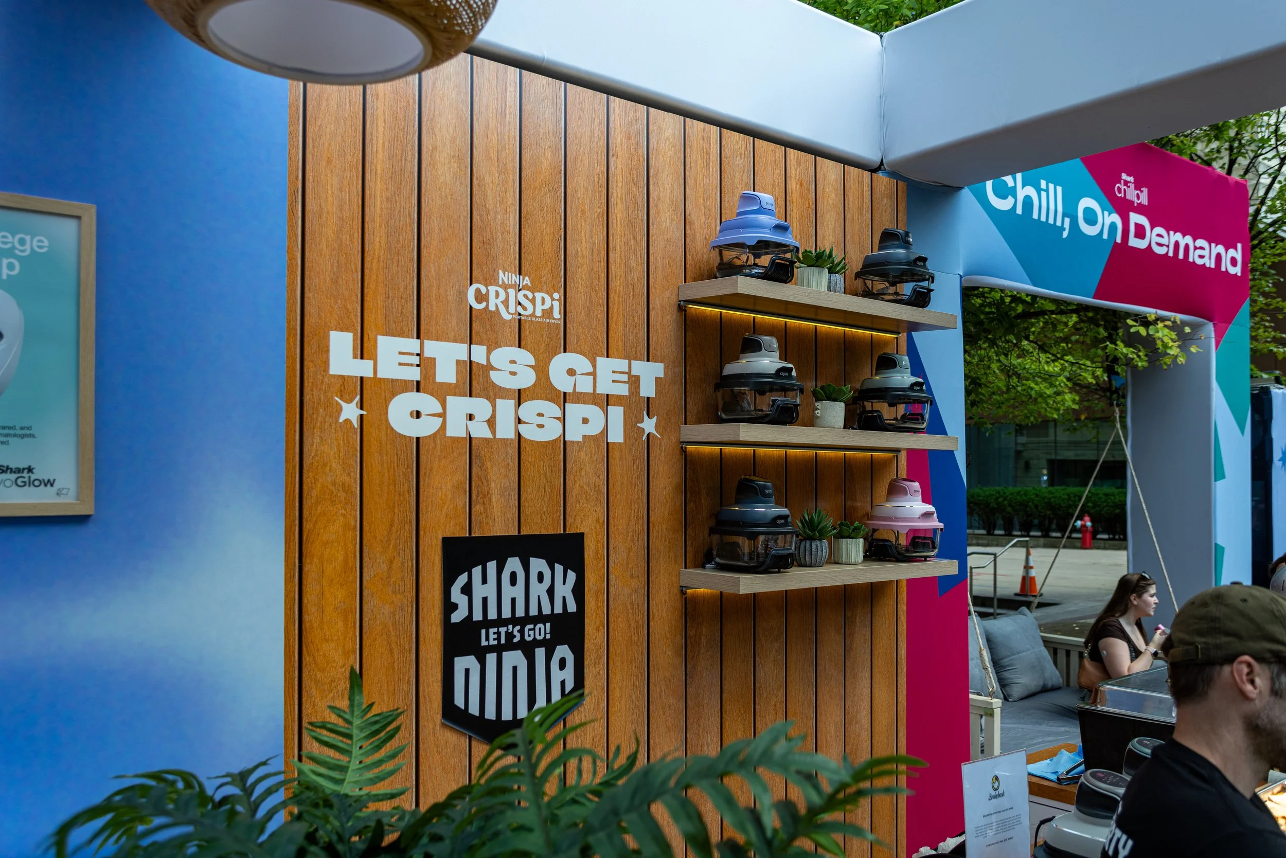 A display booth with the text 'Ninja CRISPI' and 'Let's Get Crispy' on a wooden wall, showing colorful air fryers on shelves, with people sitting nearby at an outdoor event.