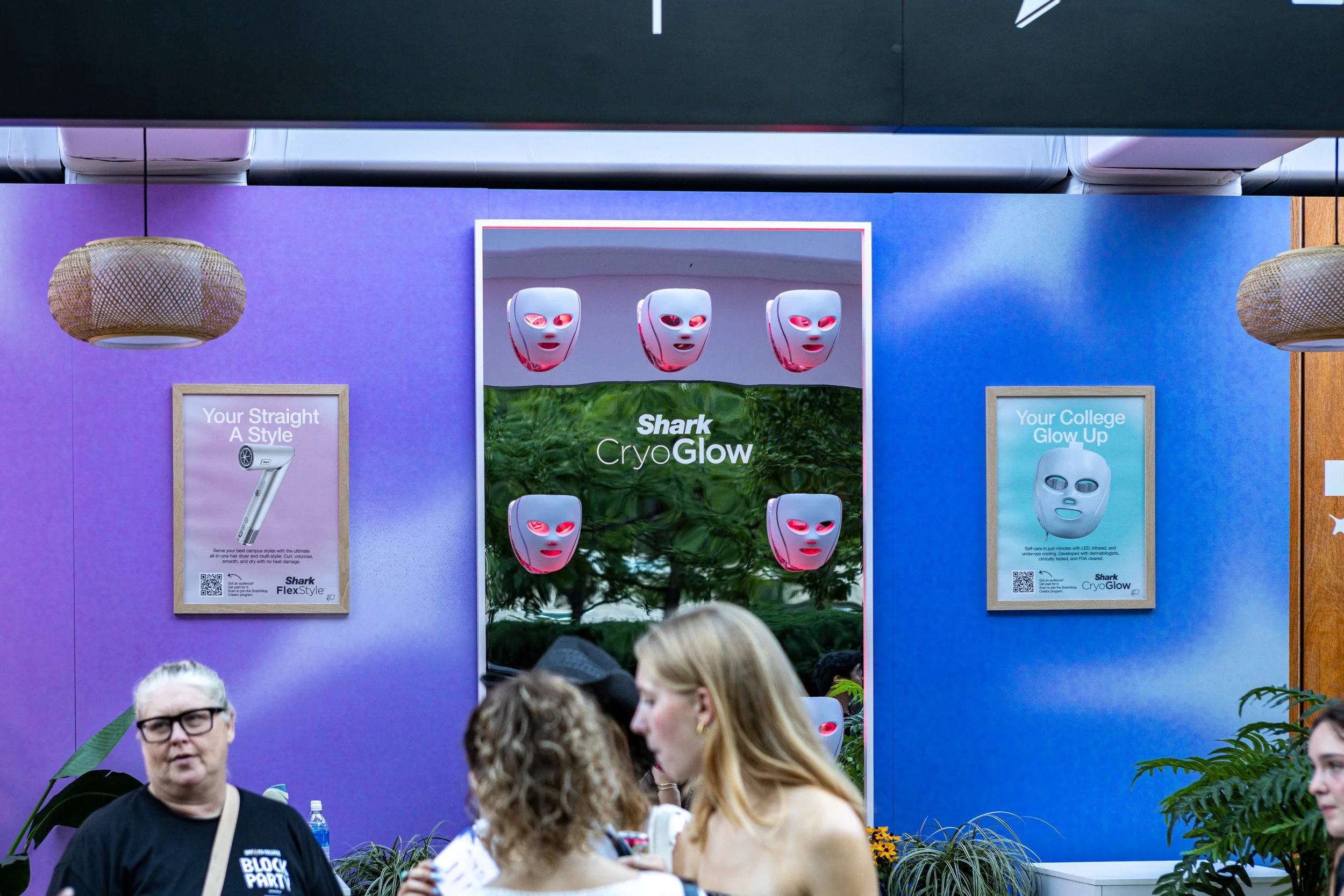 People at an indoor booth showcasing Shark CryoGlow masks, with promotional posters and purple and blue walls.