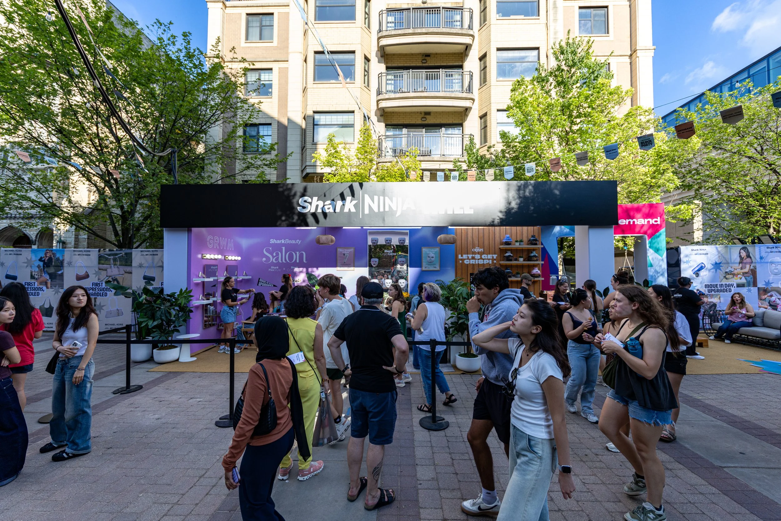A crowd of people gathered outdoors at an event with branded booths and signage, with a tall building and green trees in the background.