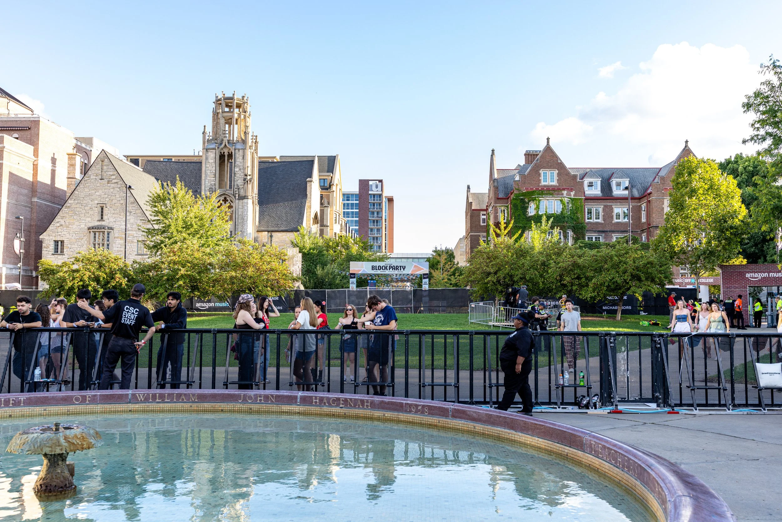People gathered at a fence around a fountain in an urban park with historic and modern buildings in the background, and a clear blue sky overhead.