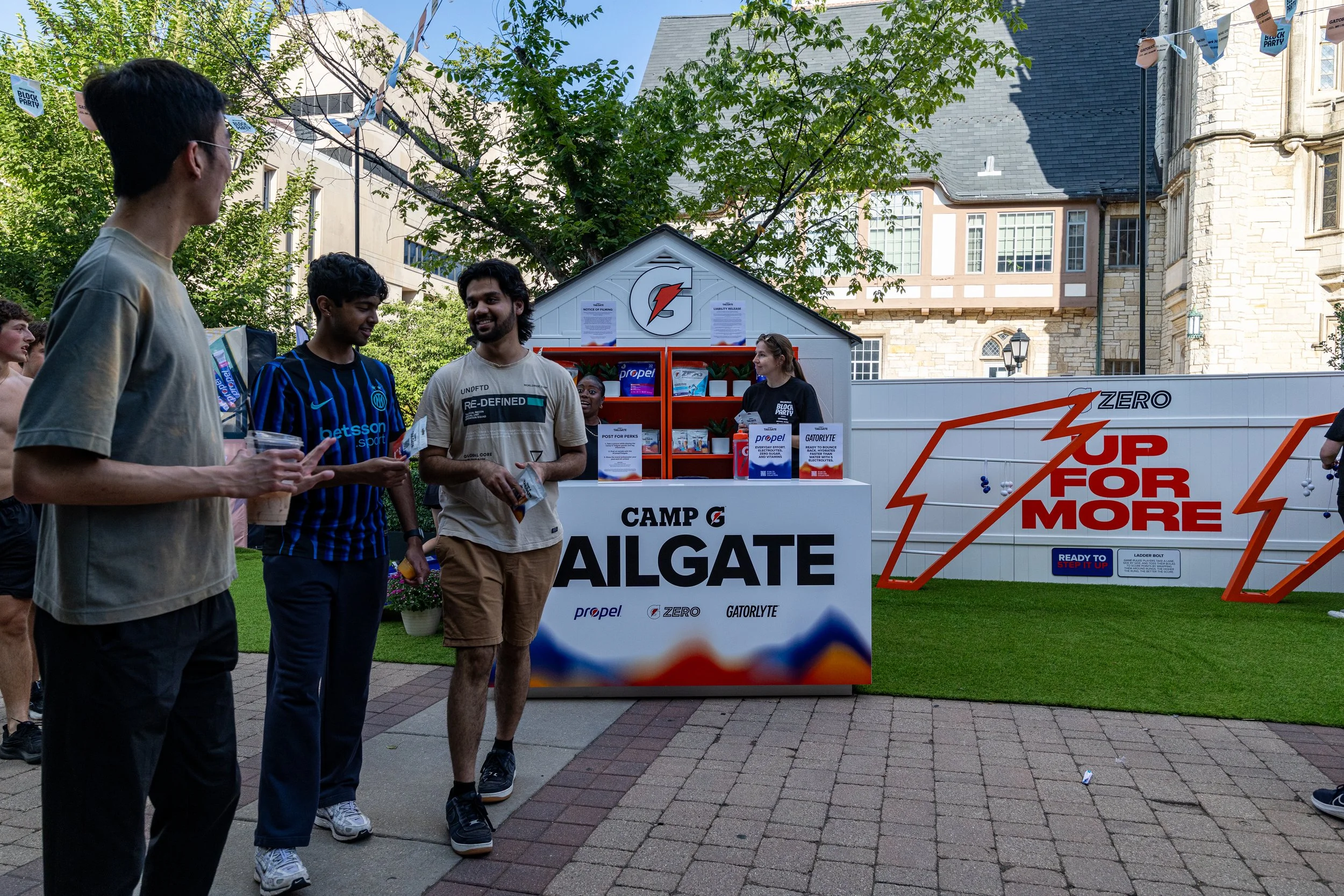 A group of young adults outdoors at a community event, engaging in conversation. Behind them is a white booth with signs promoting dental hygiene products and a large sign with the words 'Camp Algate' and slogans like 'Zero Up for More'. There are tr