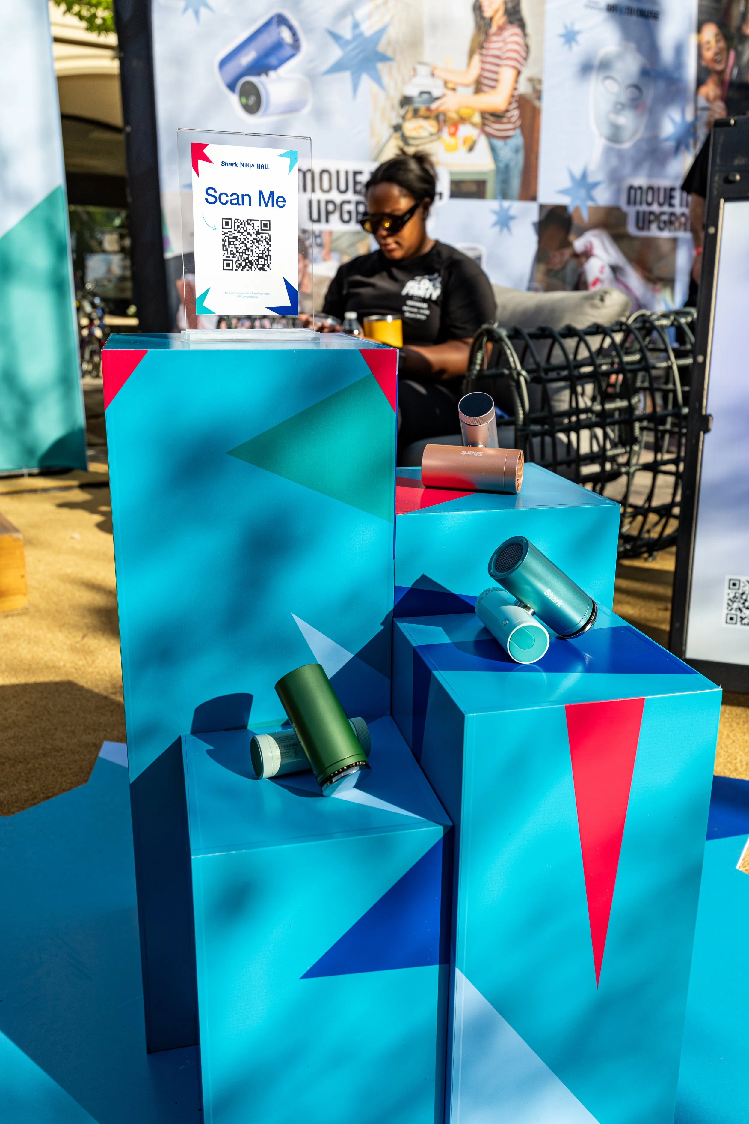 Display booth with Shark and Ninja branded portable hair dryers on blue and red angular platforms. Sign with QR code reads 'Scan Me'.
