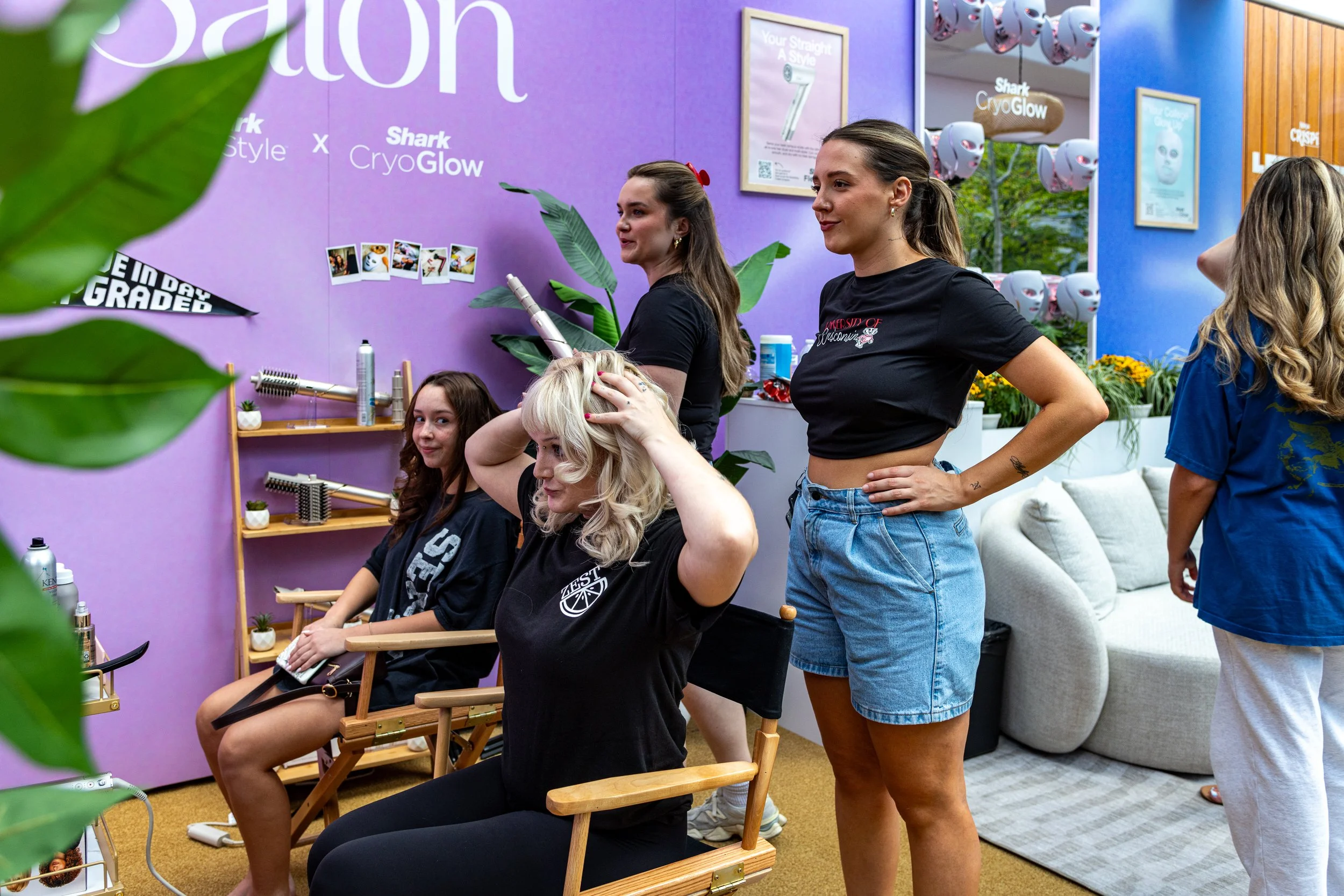 Women sitting in chairs getting hair styling treatment at a salon or event with a purple wall, promotional posters, and hanging hair drying caps in the background.
