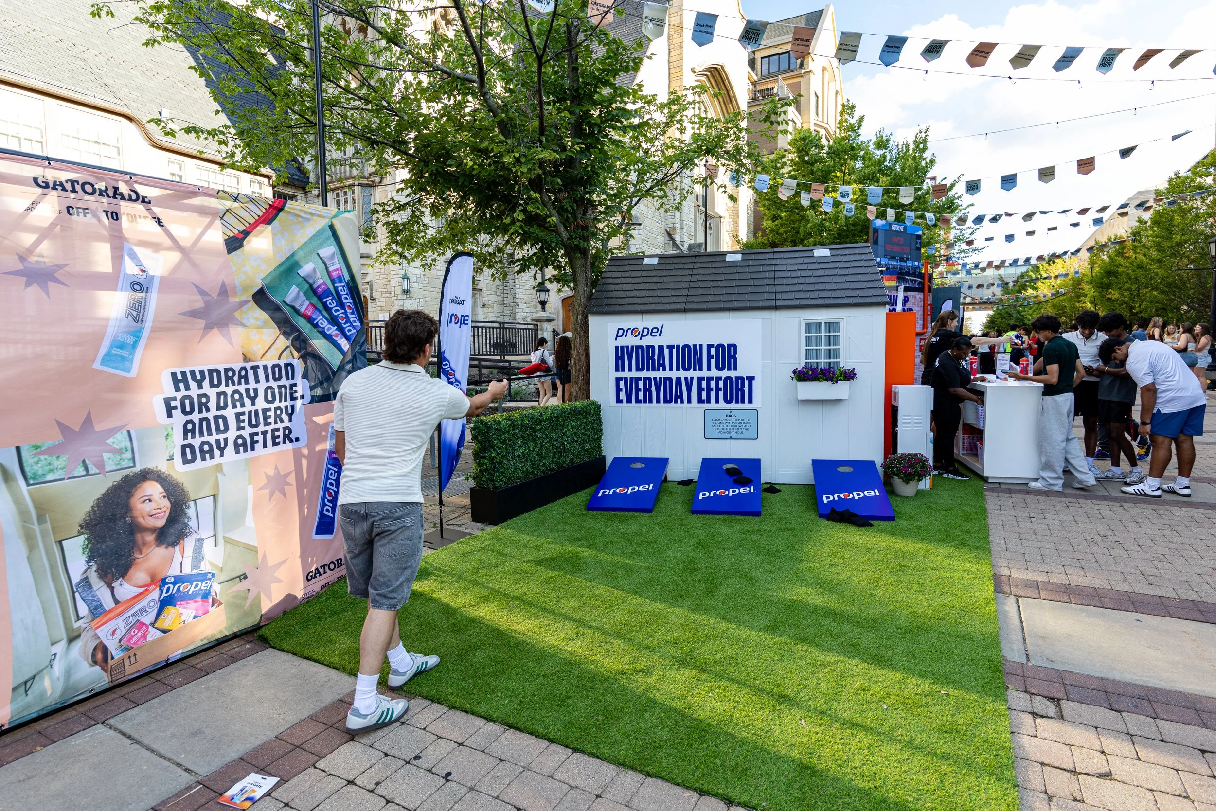 People attending an outdoor promotional event for Propel hydration products, with branded banners, a small house-like display, and overhead string flags, on a sunny day in a city setting with trees and buildings in the background.