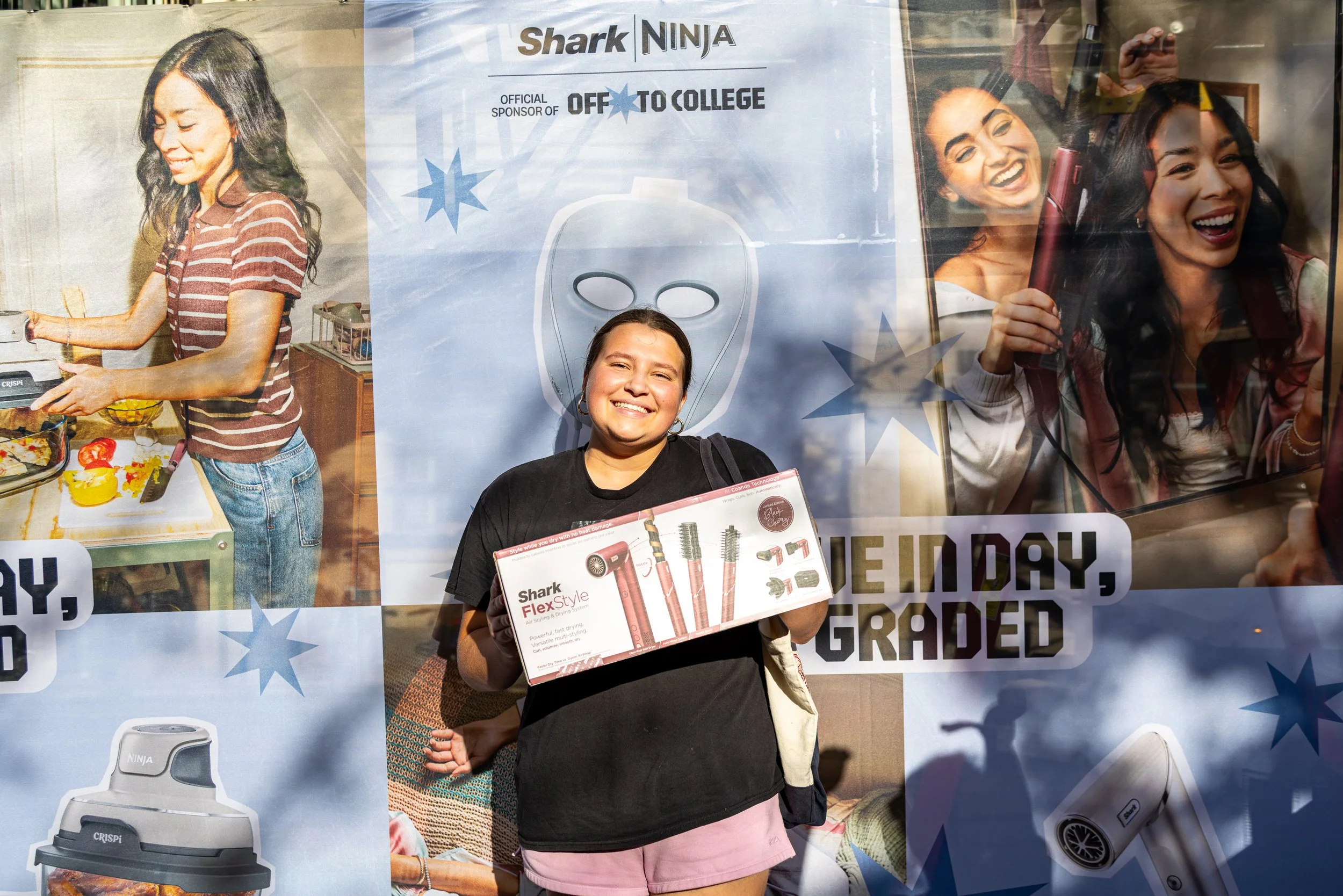 A young girl standing in front of a promotional poster, smiling, holding a Shark FlexStyle hair styling device box. The poster features images of women using hair styling tools and has text referring to Shark and off to college.