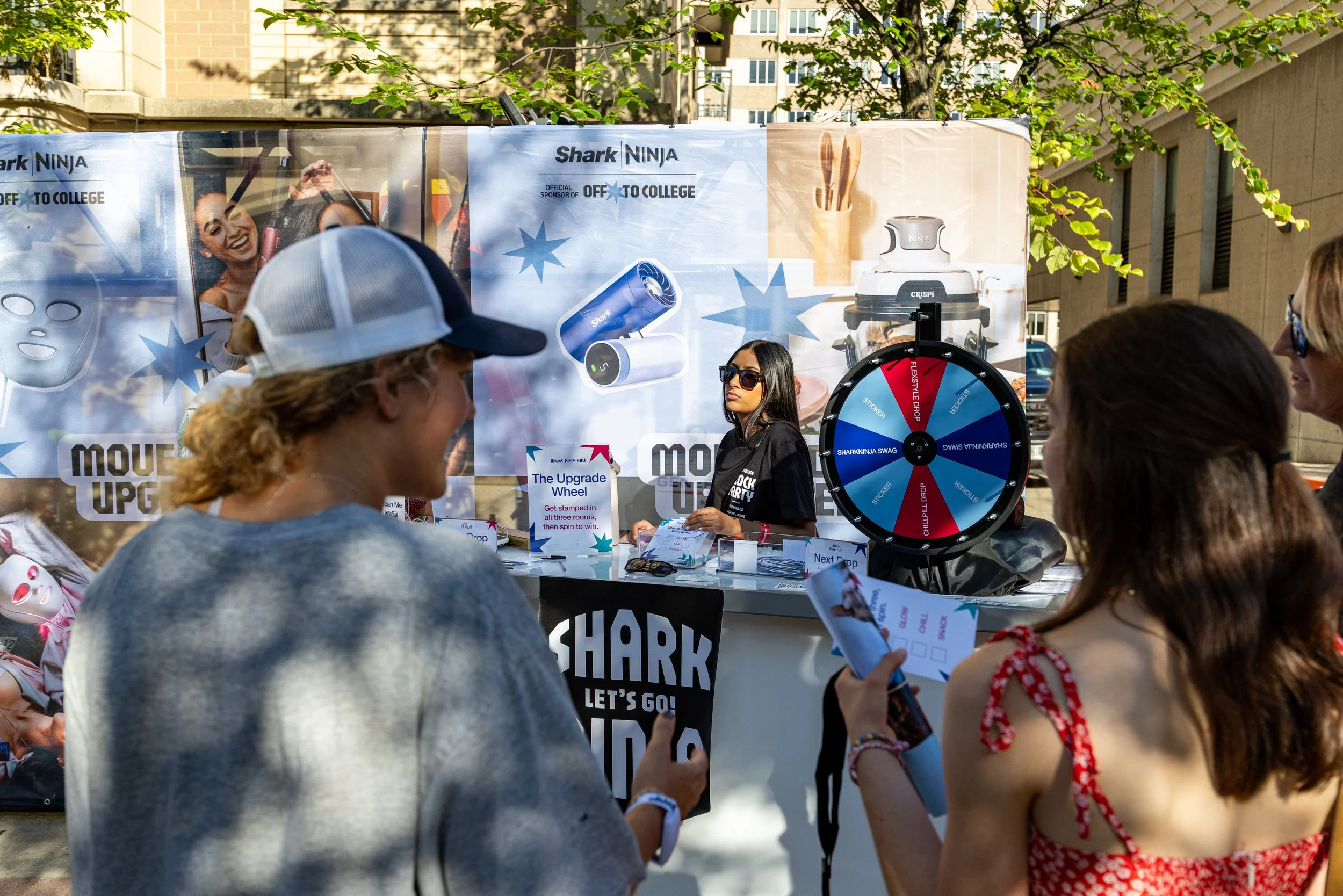 Several people gathered outdoors at a promotional event with a Shark vacuum cleaner on display. A woman stands behind a table, and a spinning wheel is nearby. A large banner behind them features images of a smiling woman and Shark products. The scene