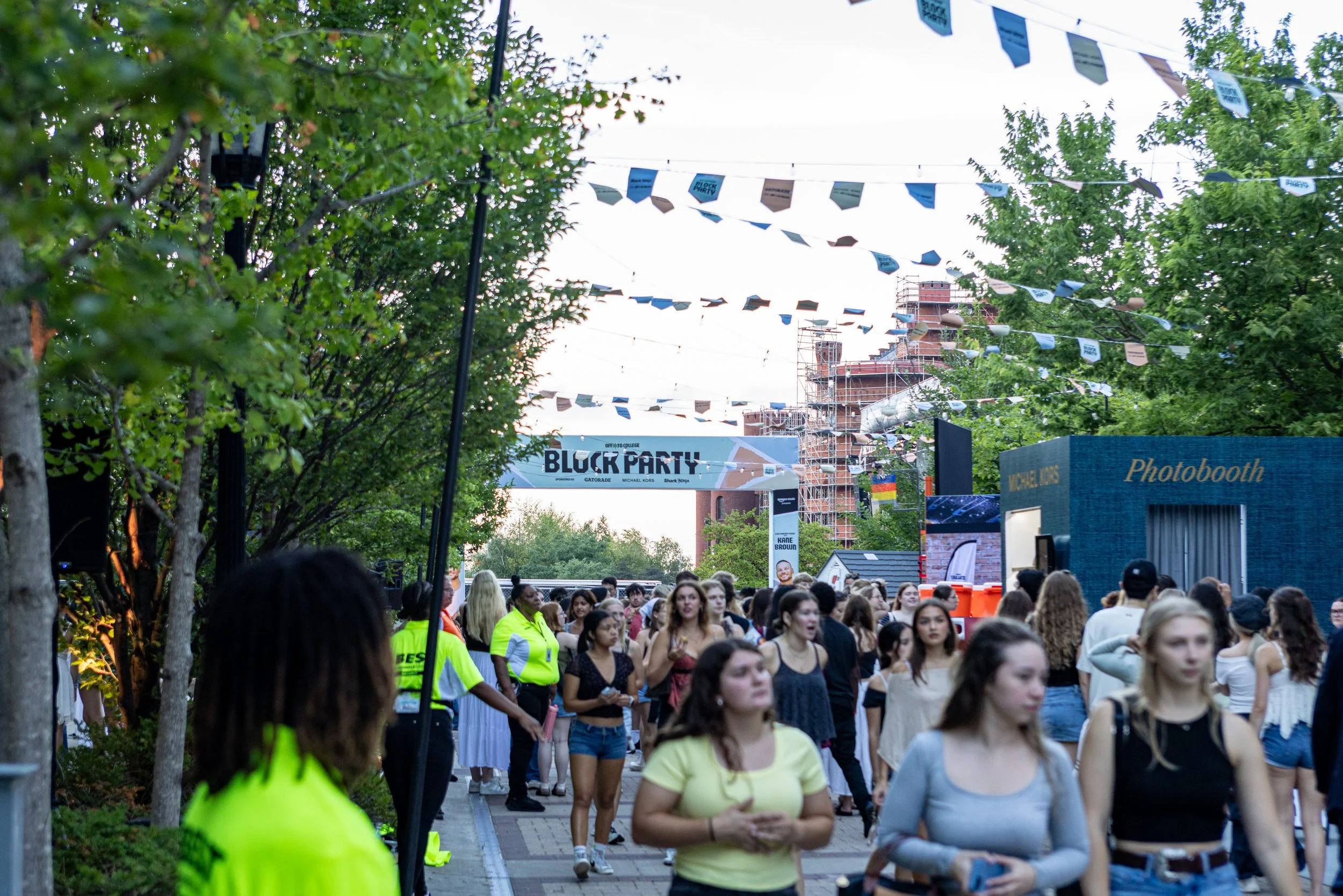 A crowd of people walking outdoors during a festival. There are trees and string banners overhead, and a building in the background under construction. A sign reads 'BLOCK PARTY'.