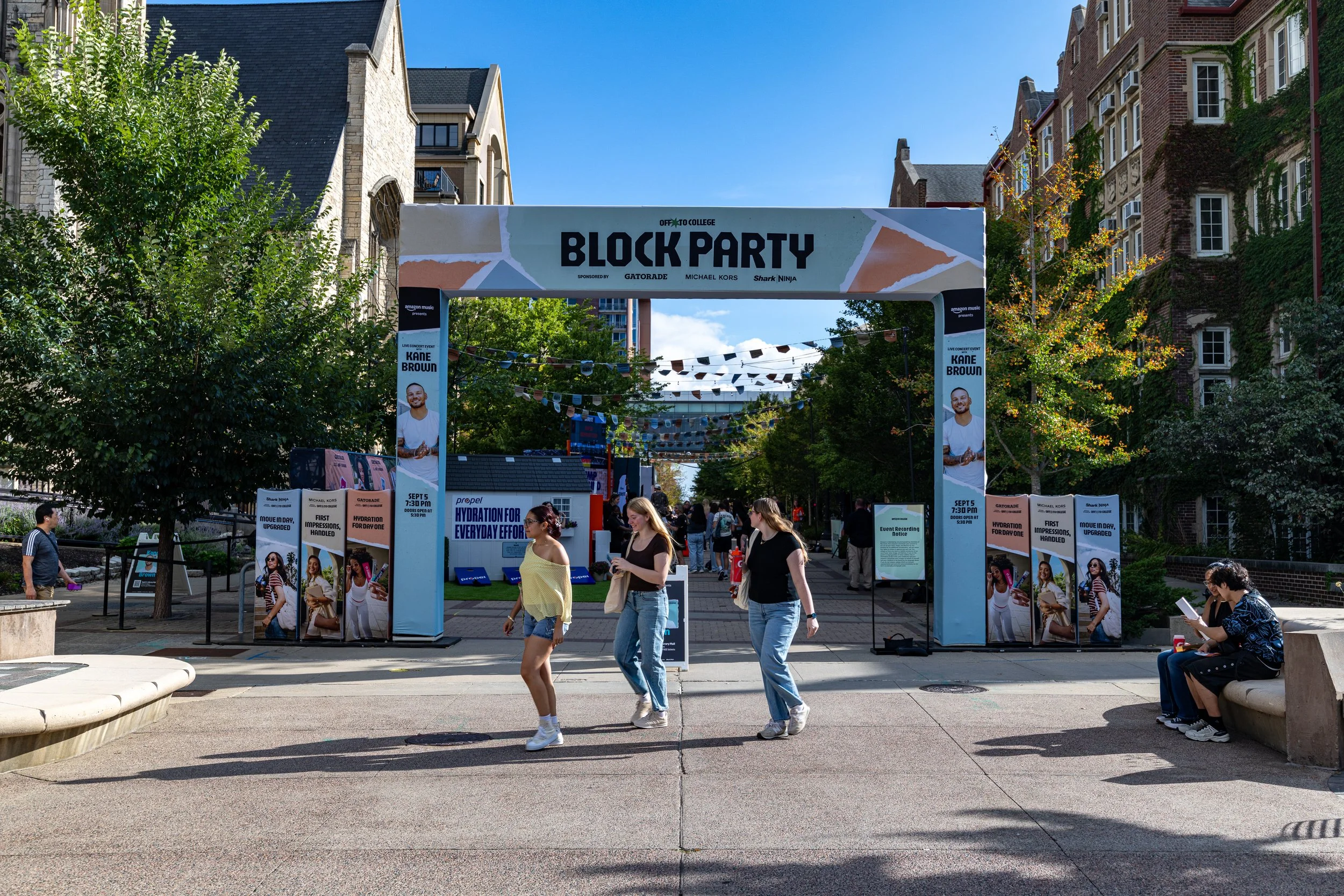 People walking and sitting in a park-like area during a block party with tents, posters, and string lights, surrounded by trees and brick buildings.