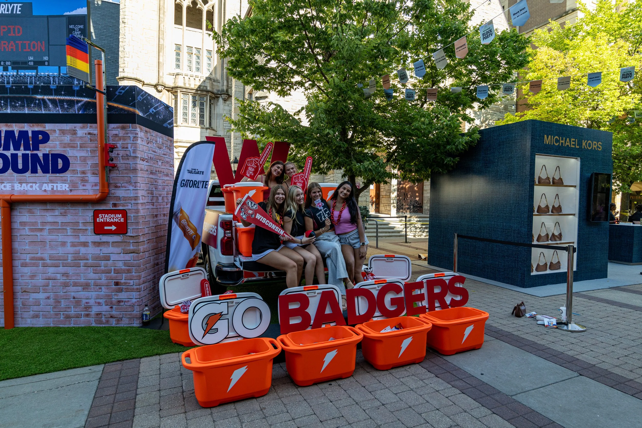 Group of five young women sitting inside an orange utility vehicle, smiling and holding signs with Wisconsin and Badgers logos, surrounded by orange containers labeled with sports and energy drink logos, at a street event with banners and a display o