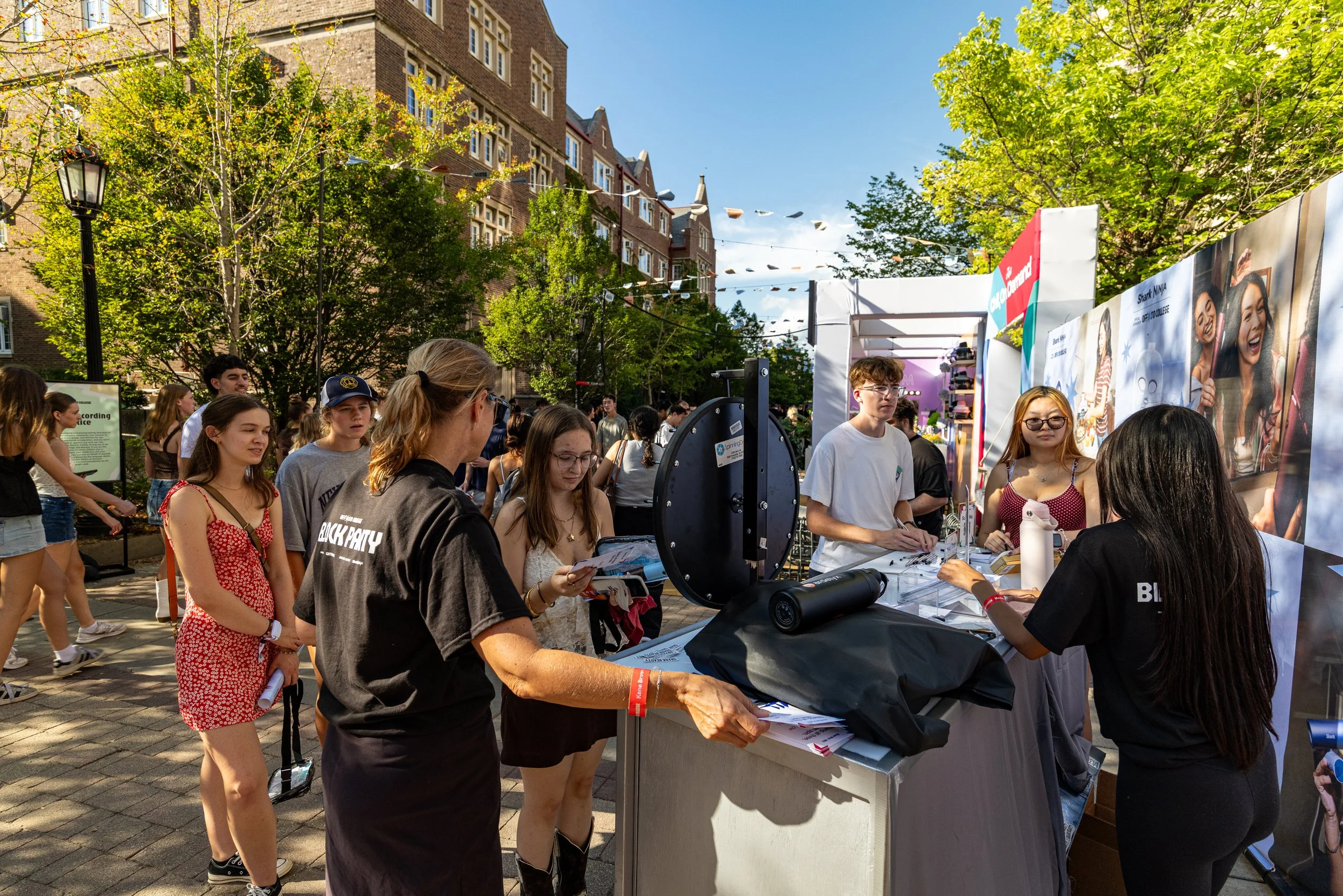 A diverse group of people gathered at an outdoor event on a sunny day, with some engaging at a booth and others walking or talking. Tall brick buildings and green trees are in the background, and there are decorative banners overhead.