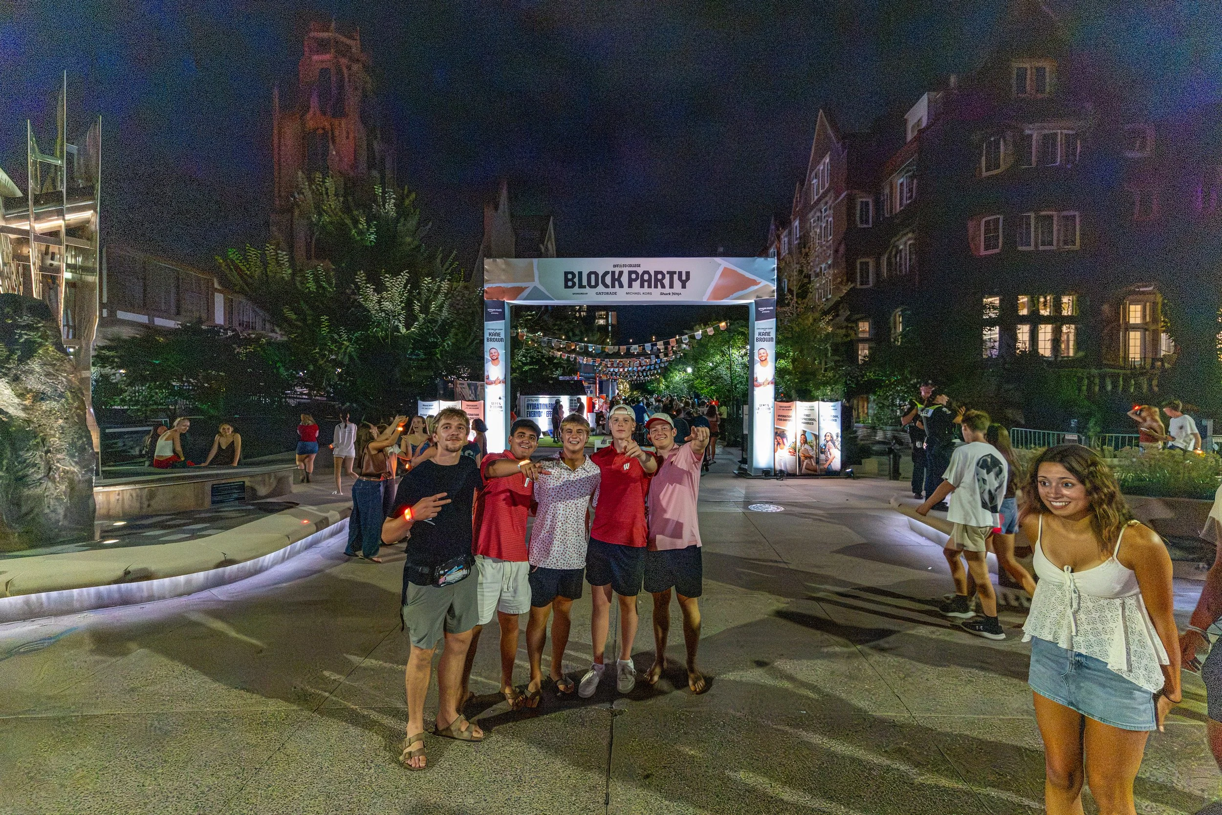 A group of young people smiling and posing for a photo at night during a block party. The scene is lively with other attendees in the background, hanging decorations, and buildings illuminated behind trees.