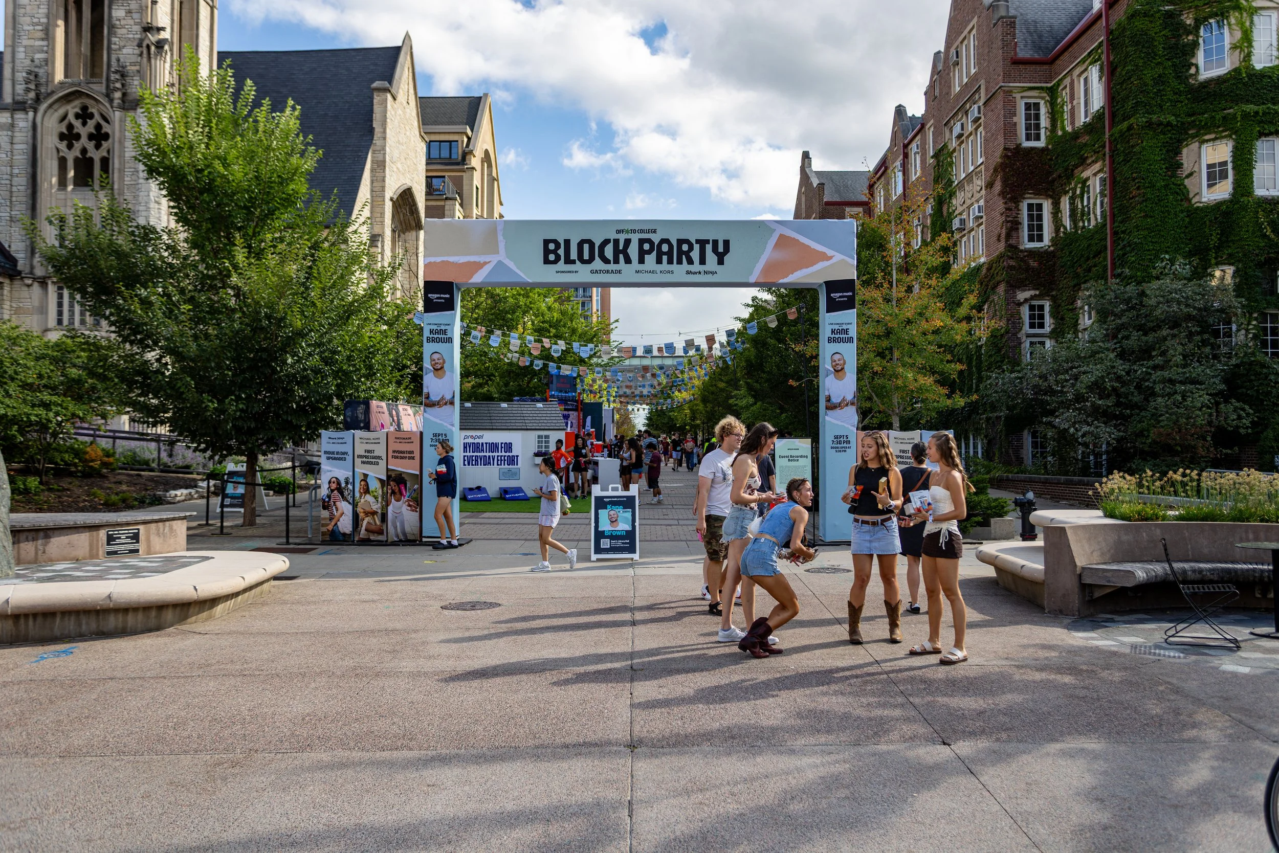 An outdoor block party on a city street with people socializing under an archway banner that reads 'Block Party'. The street is lined with tall buildings, some covered partially with greenery, and decorated with colorful flags and banners. There are 