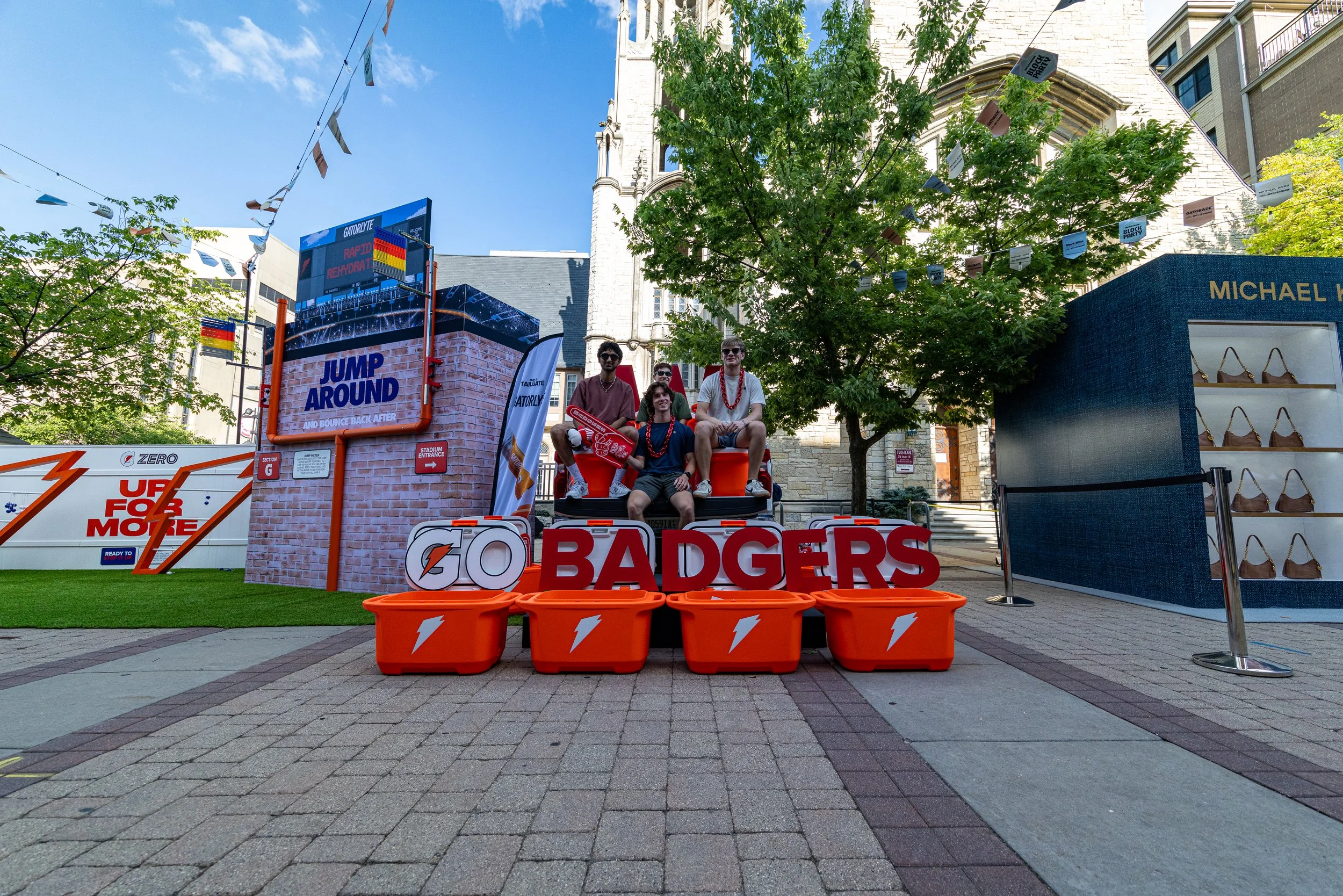Four young people sitting on a parade float with a sign that says 'BADGERS' in an outdoor plaza. The float is decorated with orange buckets and a digital screen displaying text, with banners and trees around. The background shows city buildings and a