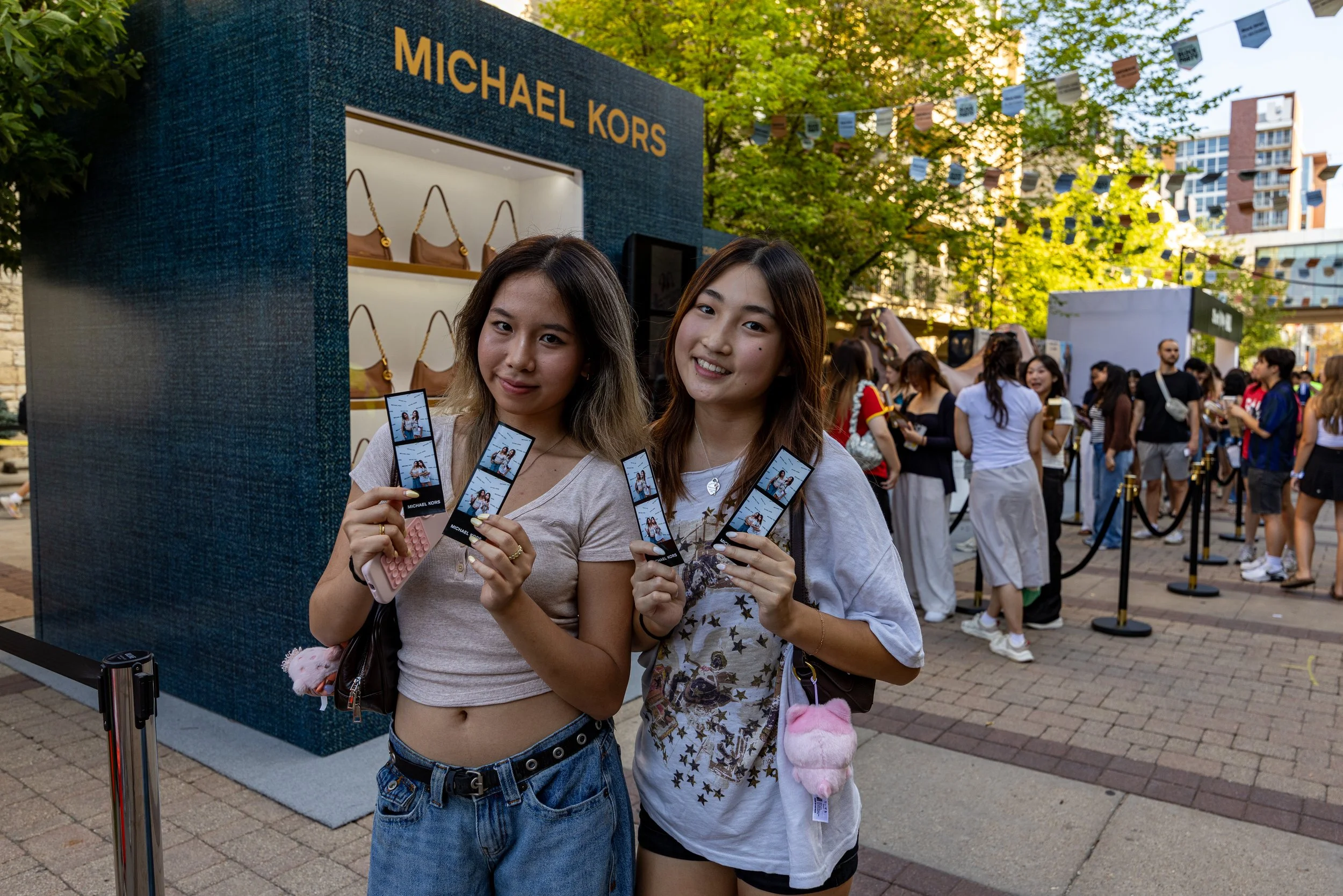Two young women are standing outdoors at what appears to be an event. They are smiling and holding small Michael Kors shopping bags. One woman is wearing a crop top and jeans, and the other is wearing a white t-shirt with star designs and black short