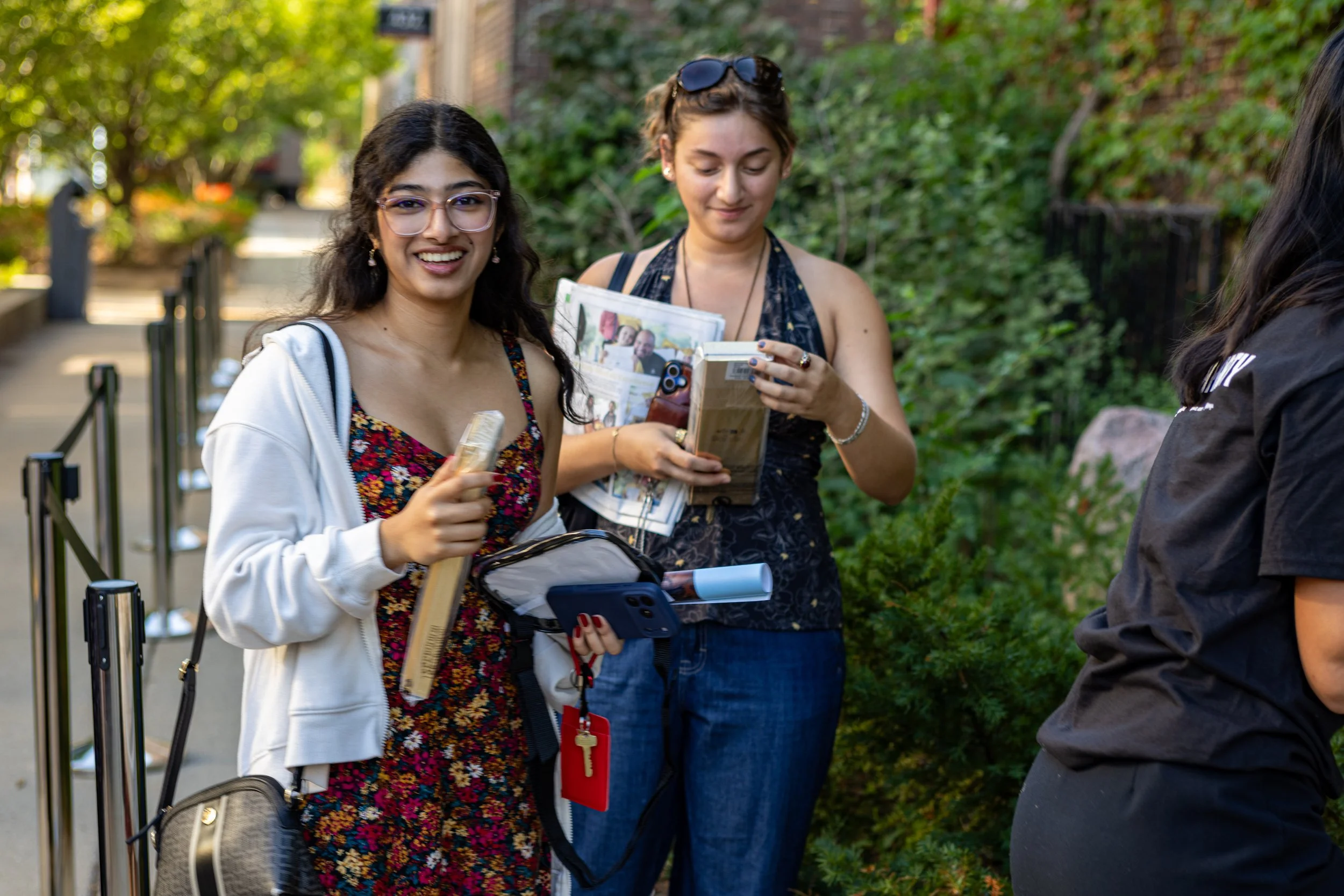 Two women standing outdoors, one smiling wearing glasses and a floral dress with a white cardigan, the other looking down at a pamphlet, both are holding items, with greenery in the background.