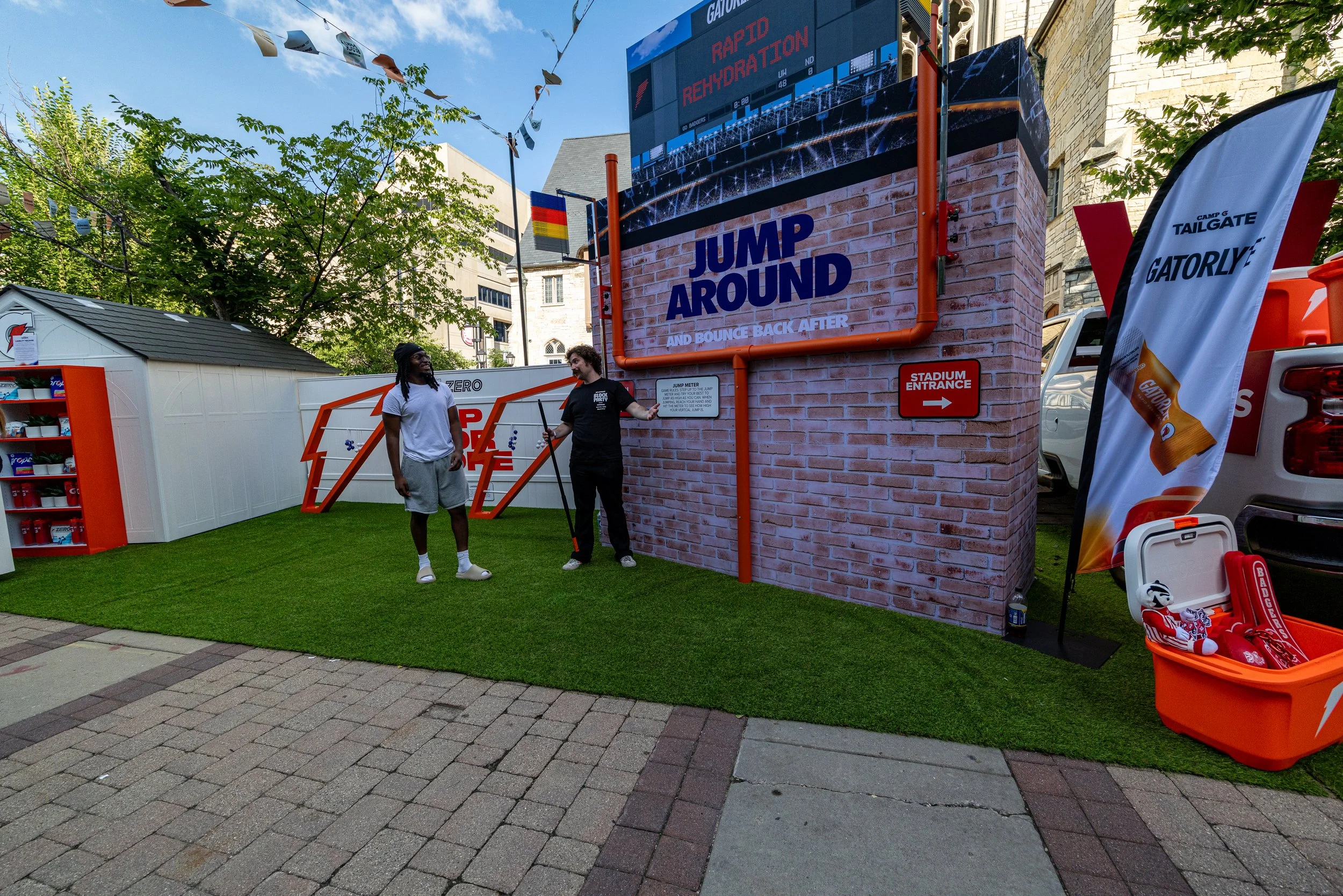 Outdoor event with a brick wall, large digital screen, and various sports-themed decorations. Two men are standing and conversing, one wearing a white t-shirt and gray shorts, and the other wearing black. There are flags, a small white building, and 