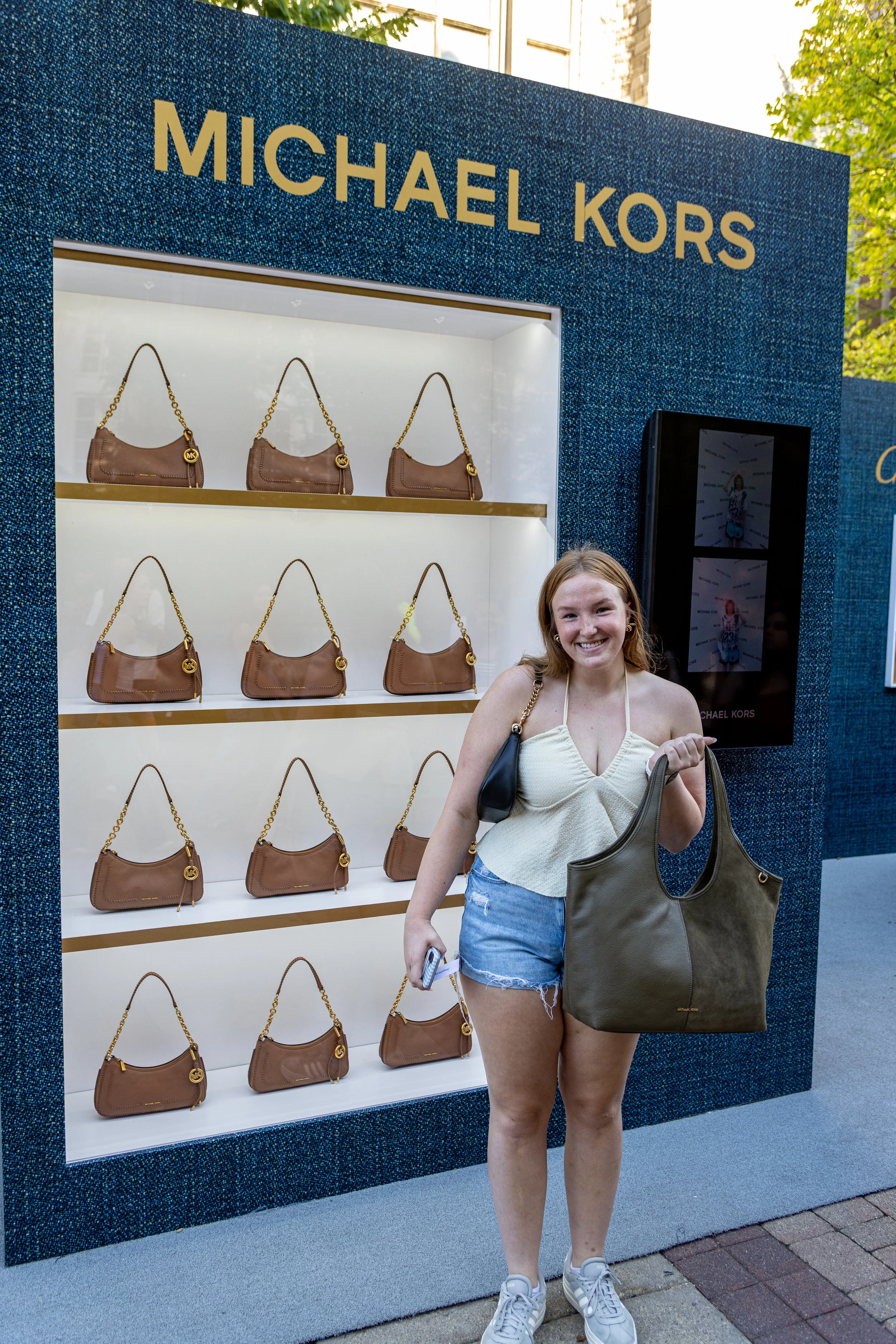 A young woman with long hair, wearing a white tank top, denim shorts, and white sneakers, stands outside in front of a display of brown Michael Kors handbags. She is smiling and holding two handbags, one in each hand.