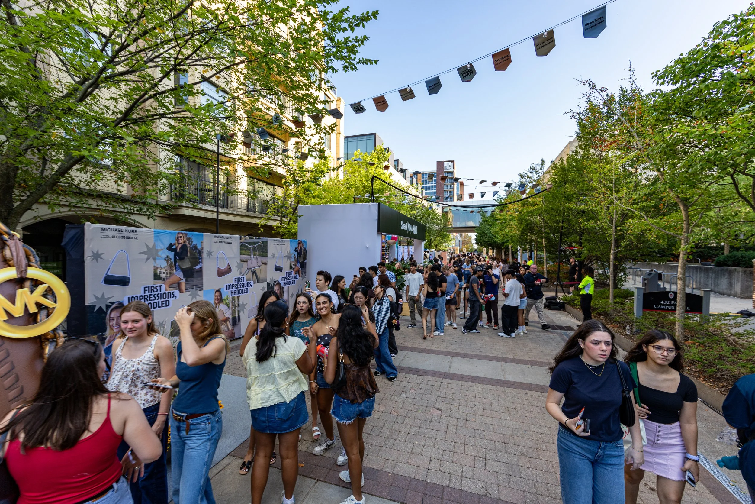 Crowd of young people walking and standing outdoor on a city street during the day, with trees, buildings, and banners strung across the street.