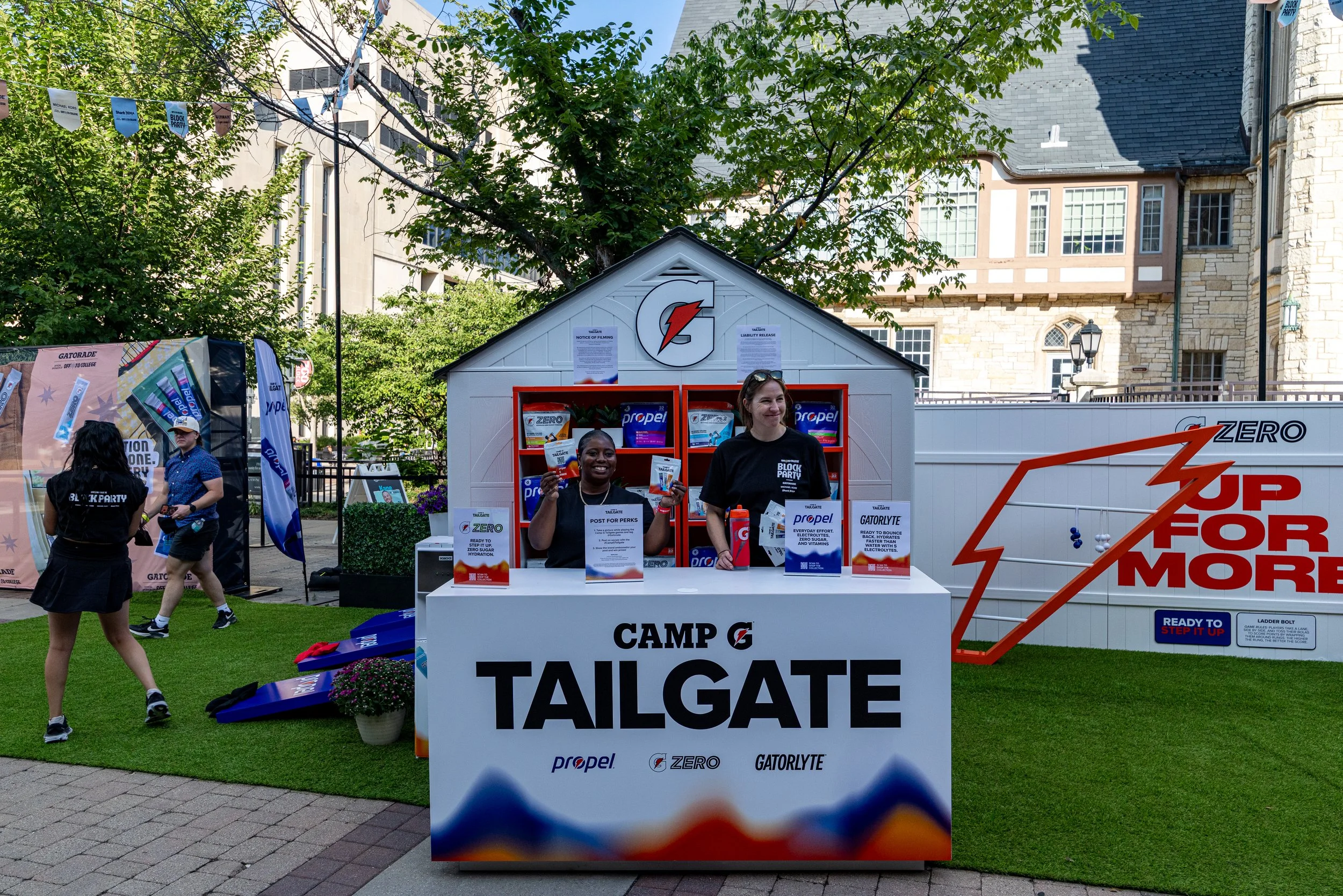 At an outdoor event, two women stand behind a white promotional booth with Gatorade branding, smiling and holding packets of Propel water flavoring. The booth has signs for Gatorade products, and the background features trees, banners, and a mix of o