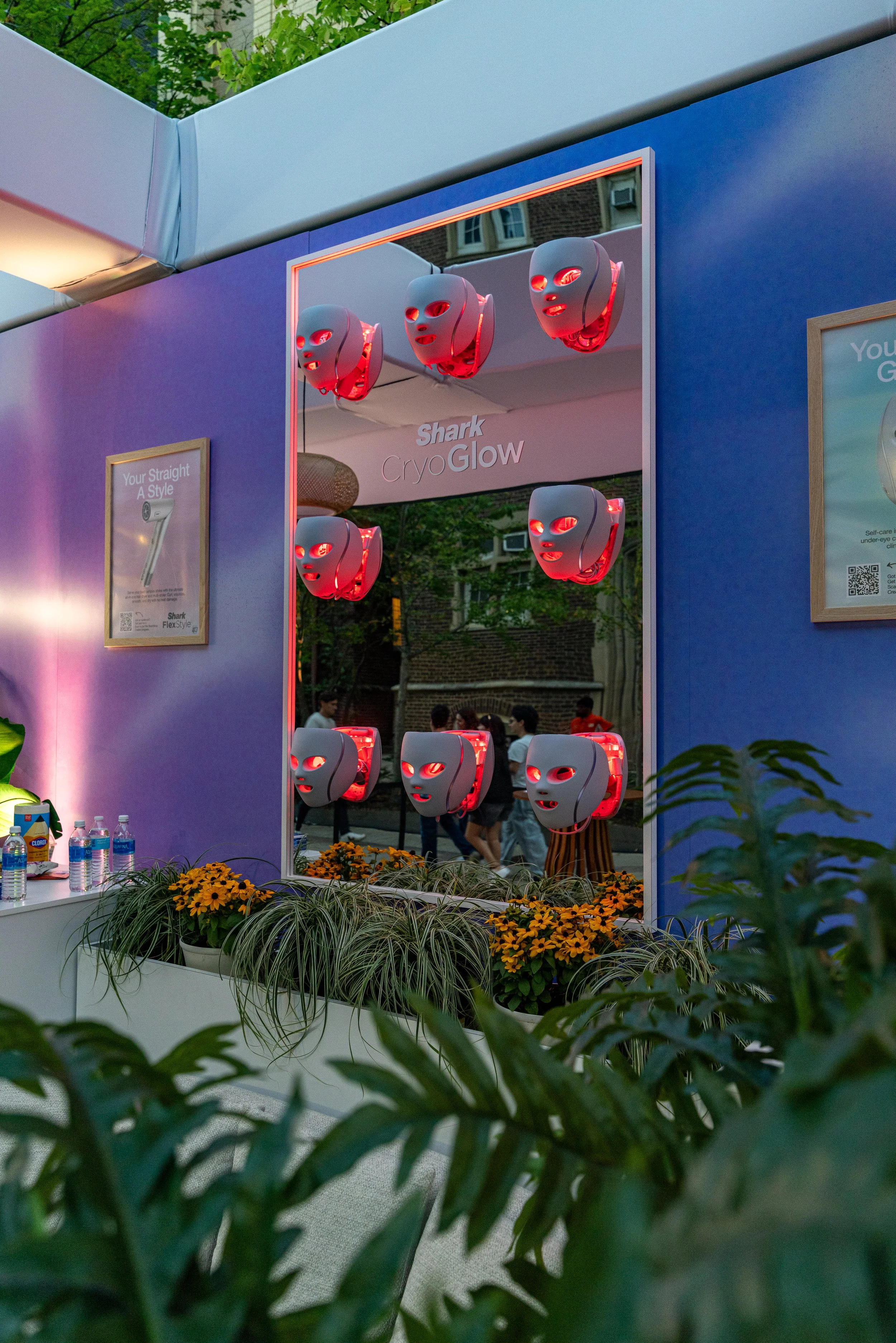 A display booth at an event showcasing Shark CryoGlow masks with multiple masks hanging on a mirror and a sign reading "Shark CryoGlow." There are potted plants, including orange flowers, in front of the booth, and water bottles on a table to the lef