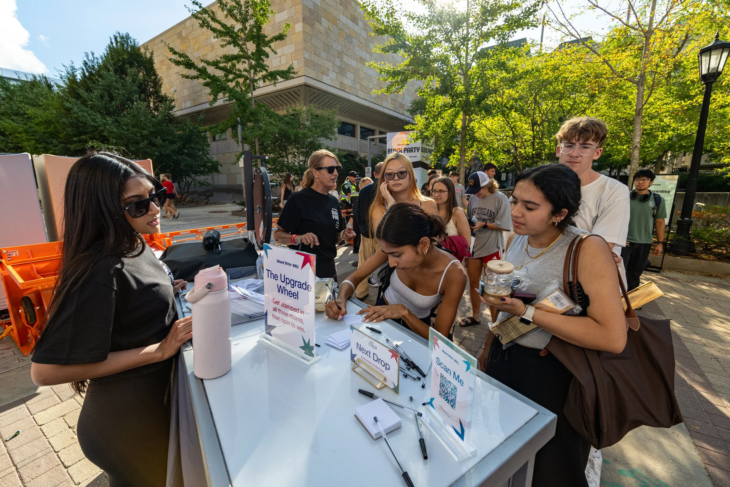 People gathered around a registration table at an outdoor event with trees and a building in the background. The table has signs, papers, and promotional items, with individuals signing in or registering.