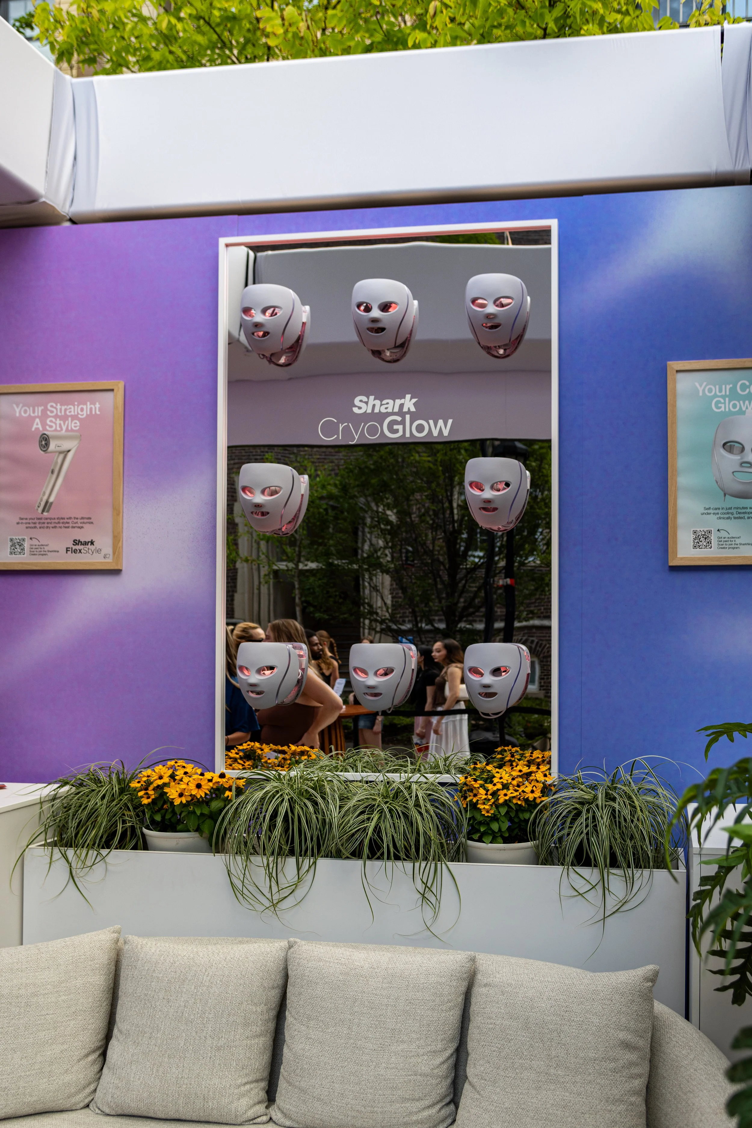Display of masks with glowing eyes and mouths at a booth named 'Shark CryoGlow,' surrounded by plants and flowers at an outdoor event.