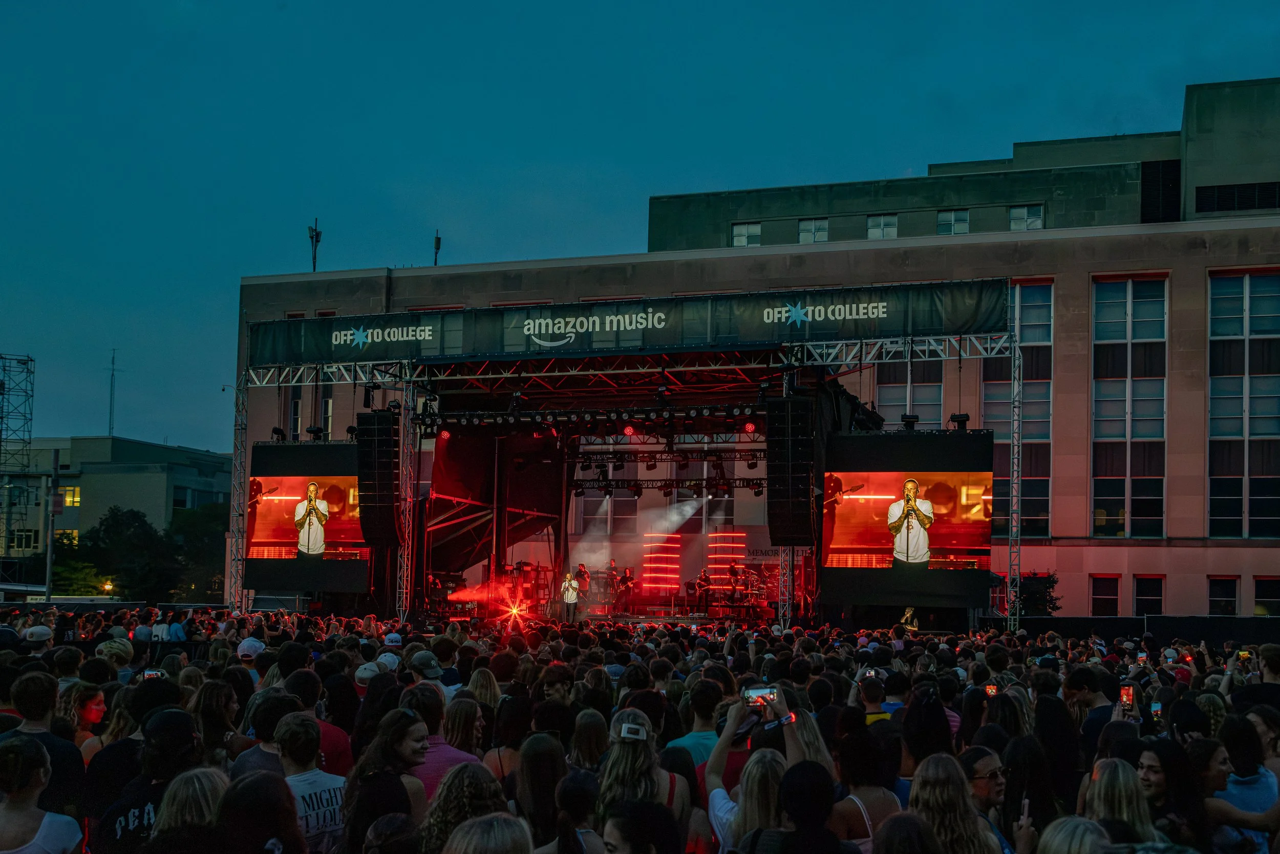 A large crowd of people watching a concert on an outdoor stage at night, with banners displaying 'Off to College' and 'Amazon Music', and performers on stage illuminated by red lights.