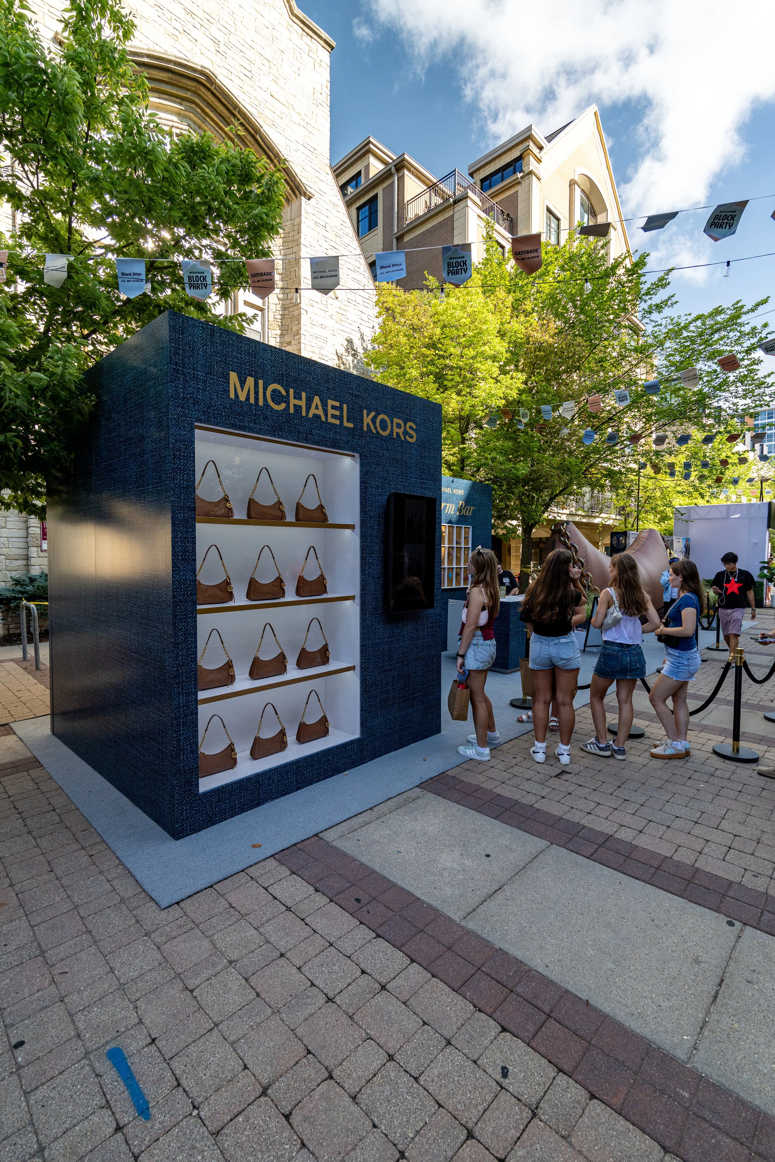 Outdoor event featuring a Michael Kors handbag display set against a building and trees with string banners that say 'BLOCK PARTY' and 'GATORADE'. People are walking and gathering around the display.