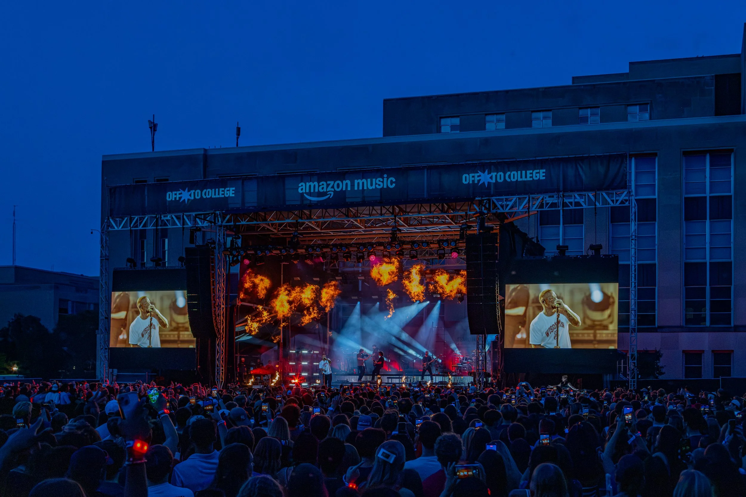 Outdoor music concert at night with a large crowd, stage with performers, flames and bright lights, and screens showing a singer.