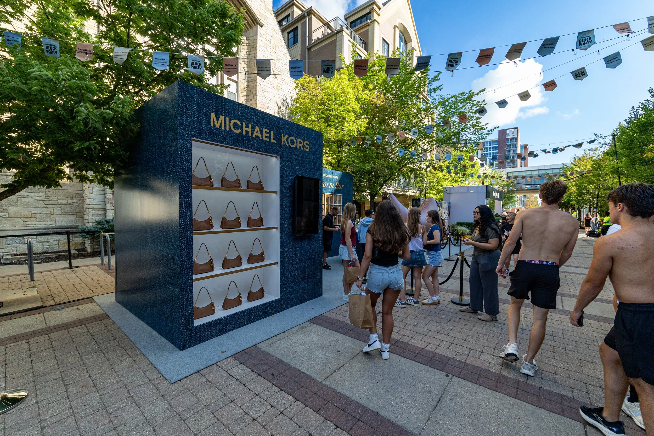 Street scene with a Michael Kors display stand showing handbags, and a group of people, some shirtless, walking and standing nearby on a pedestrian street with trees and buildings in the background, and banners hanging overhead.