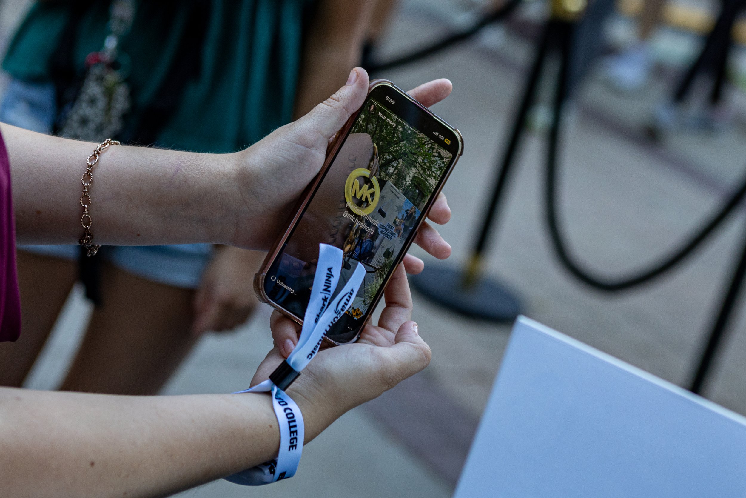 Person holding a smartphone displaying a social media story with a person in the background, wearing a bracelet and wristband.